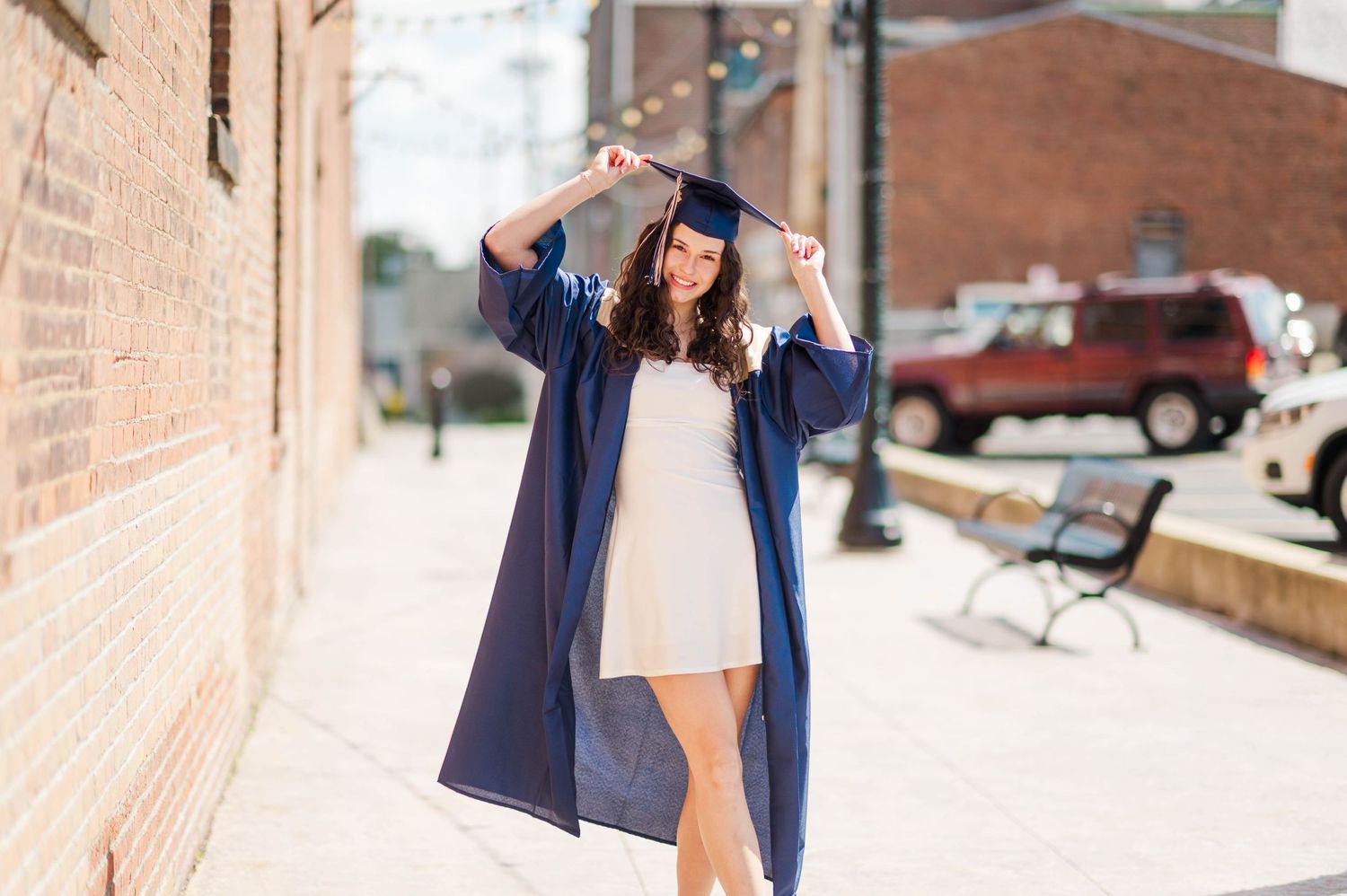 Smiling graduate in navy cap and gown adjusts her mortarboard on a sunny city sidewalk.
