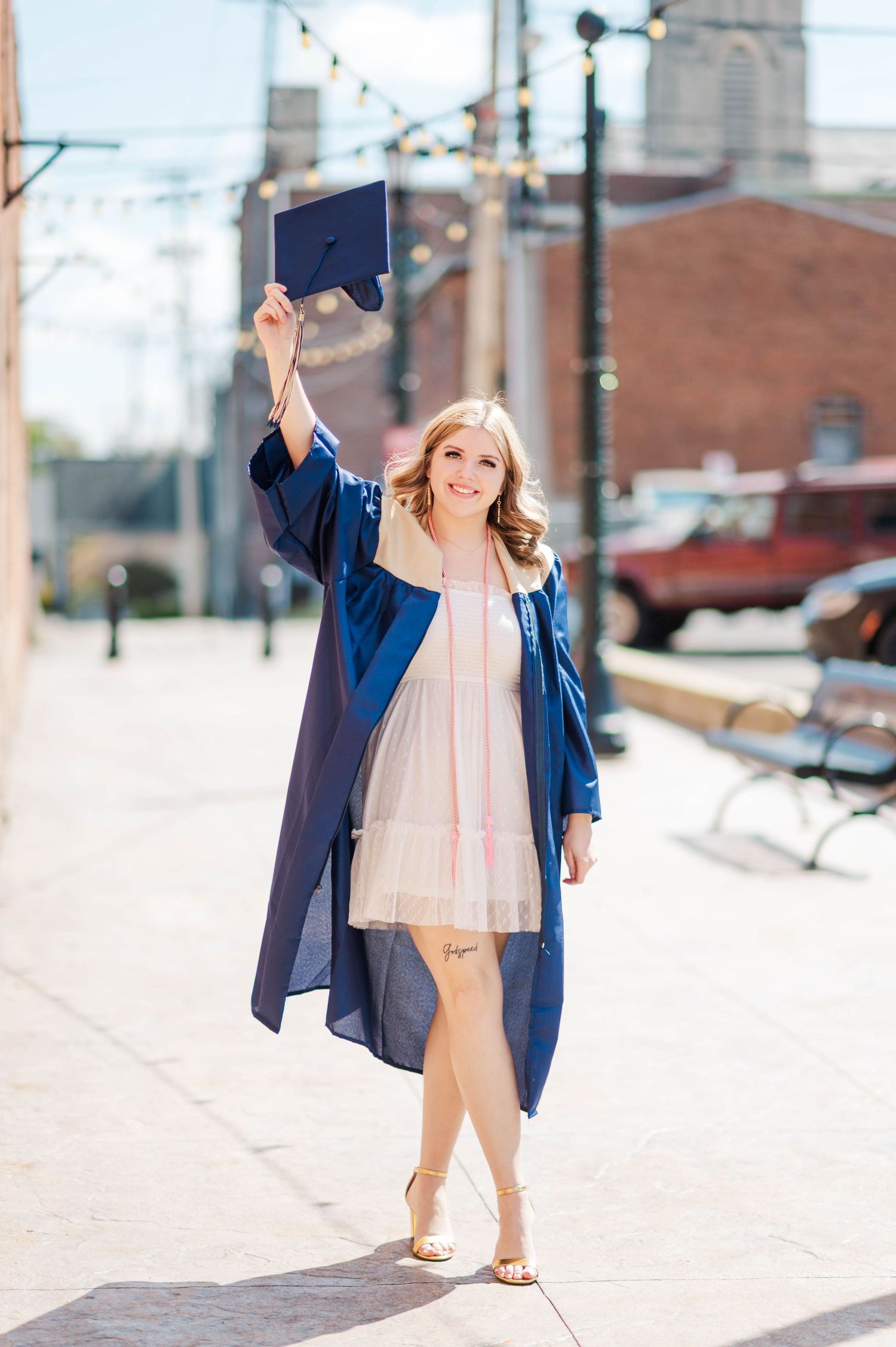 Joyful female graduate in navy gown raises mortarboard cap while walking on sunny urban sidewalk.