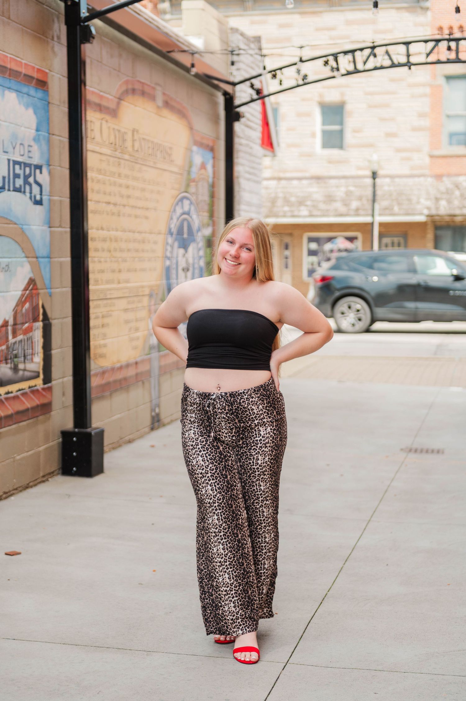 Blonde woman in black tube top and leopard print pants posing confidently on a downtown sidewalk with murals.