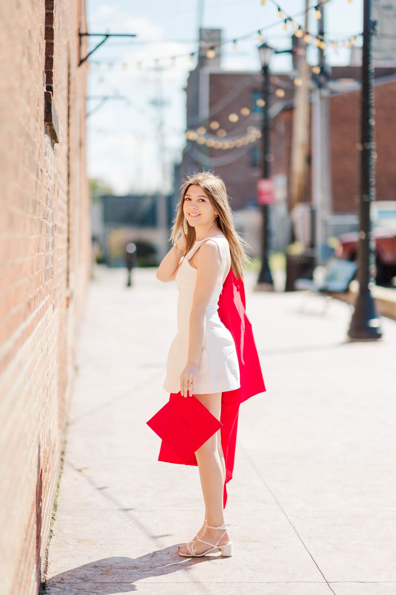 Young woman in white dress holding red graduation cap and gown on sunny urban sidewalk.