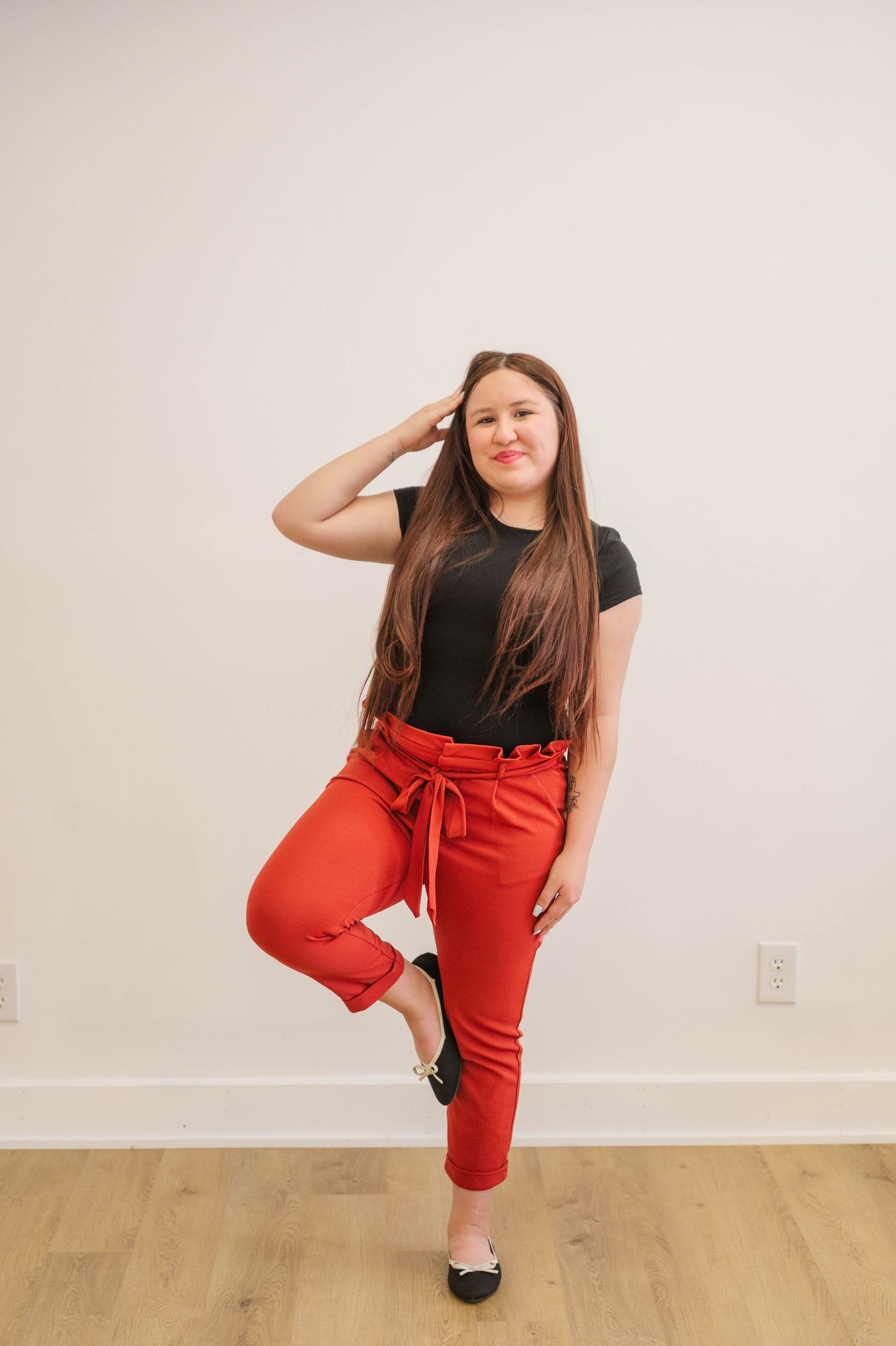 Brunette woman in black tee and red paperbag pants posing on one leg in a bright studio.