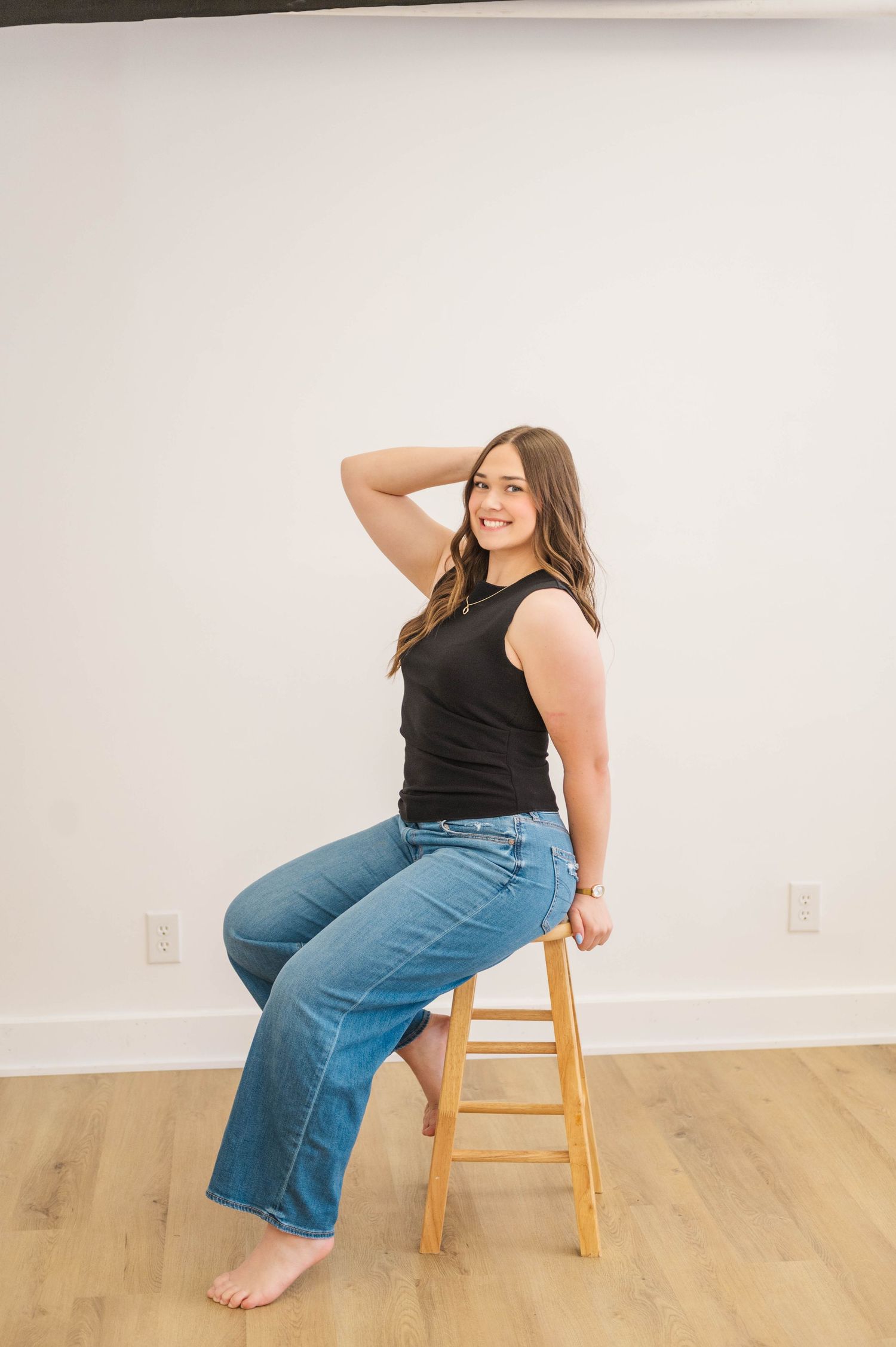 Young woman in black sleeveless top and flare jeans posing on wooden stool, smiling.