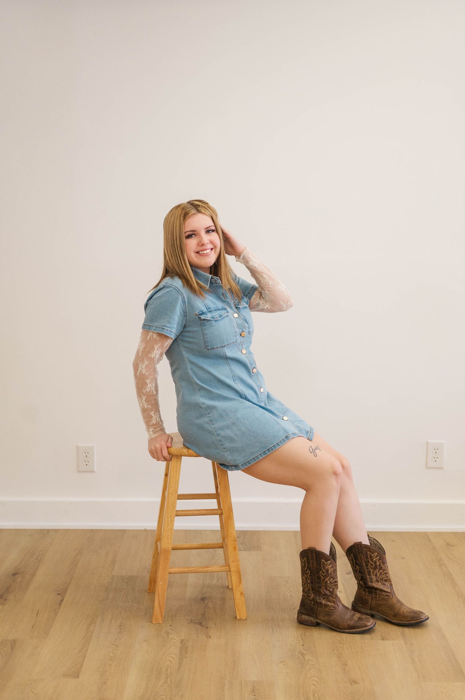 Blonde woman with tattoos sits on wooden stool in denim dress and cowboy boots, smiling confidently.