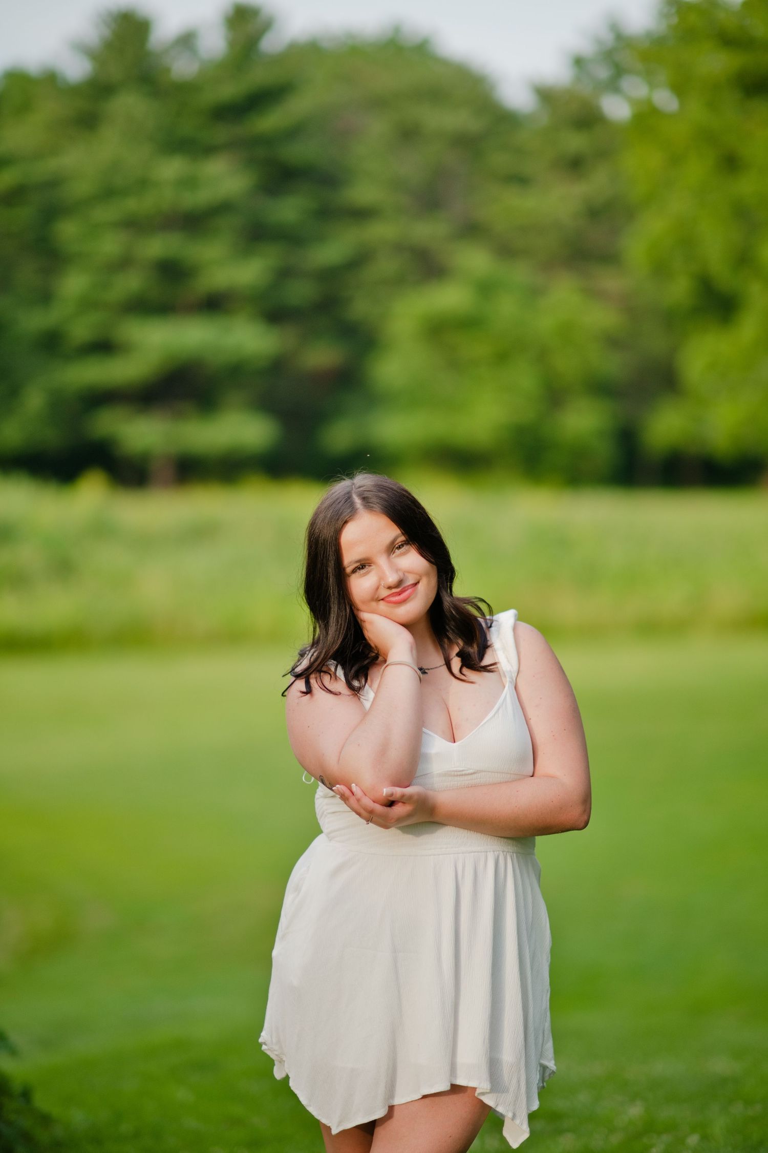 Young woman in white dress posing outdoors on green grass with lush trees in background.
