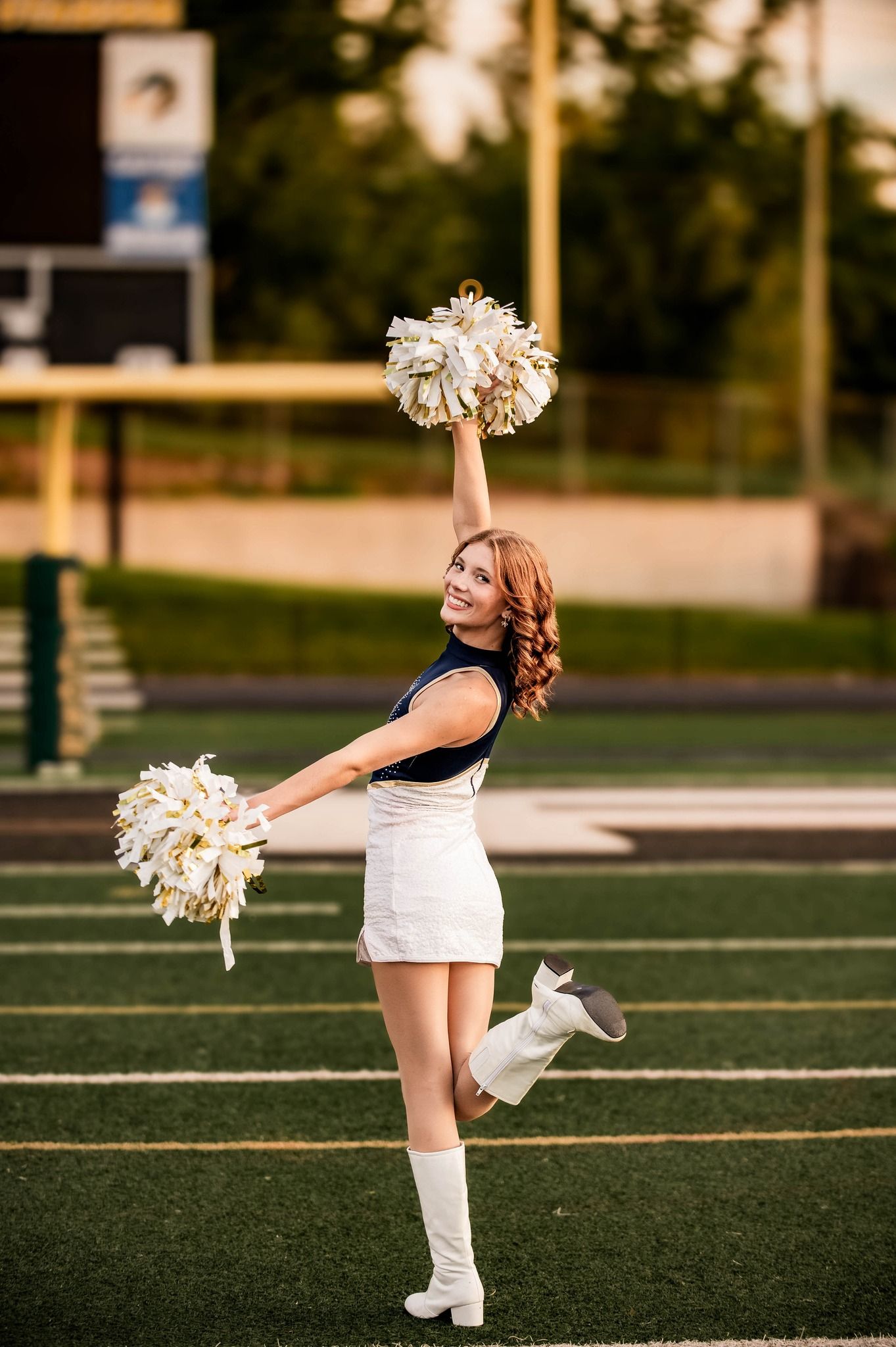 Cheerleader with pom poms striking a pose on a sports field at sunset.