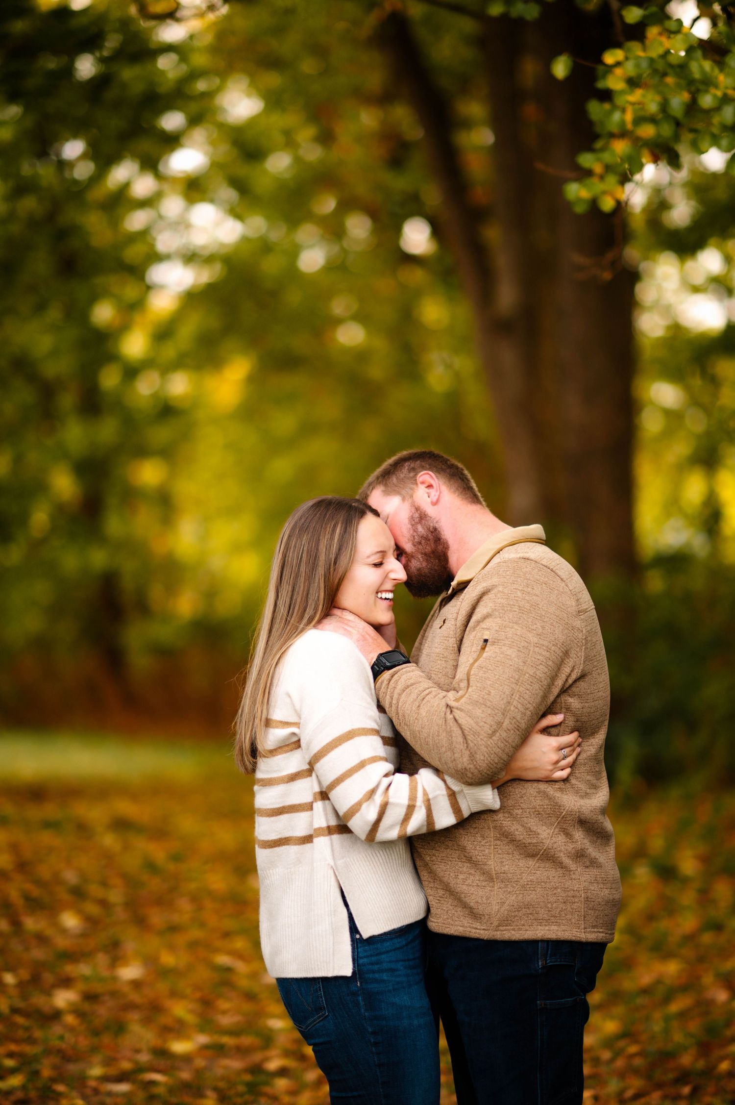 A romantic couple embraces among fall leaves in a park setting during sunset.