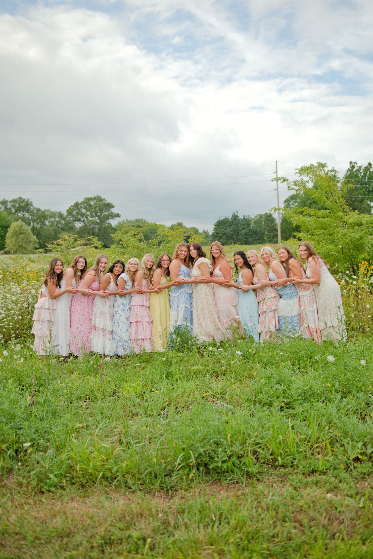 A group of young women in colorful pastel dresses pose together in a lush green meadow field.