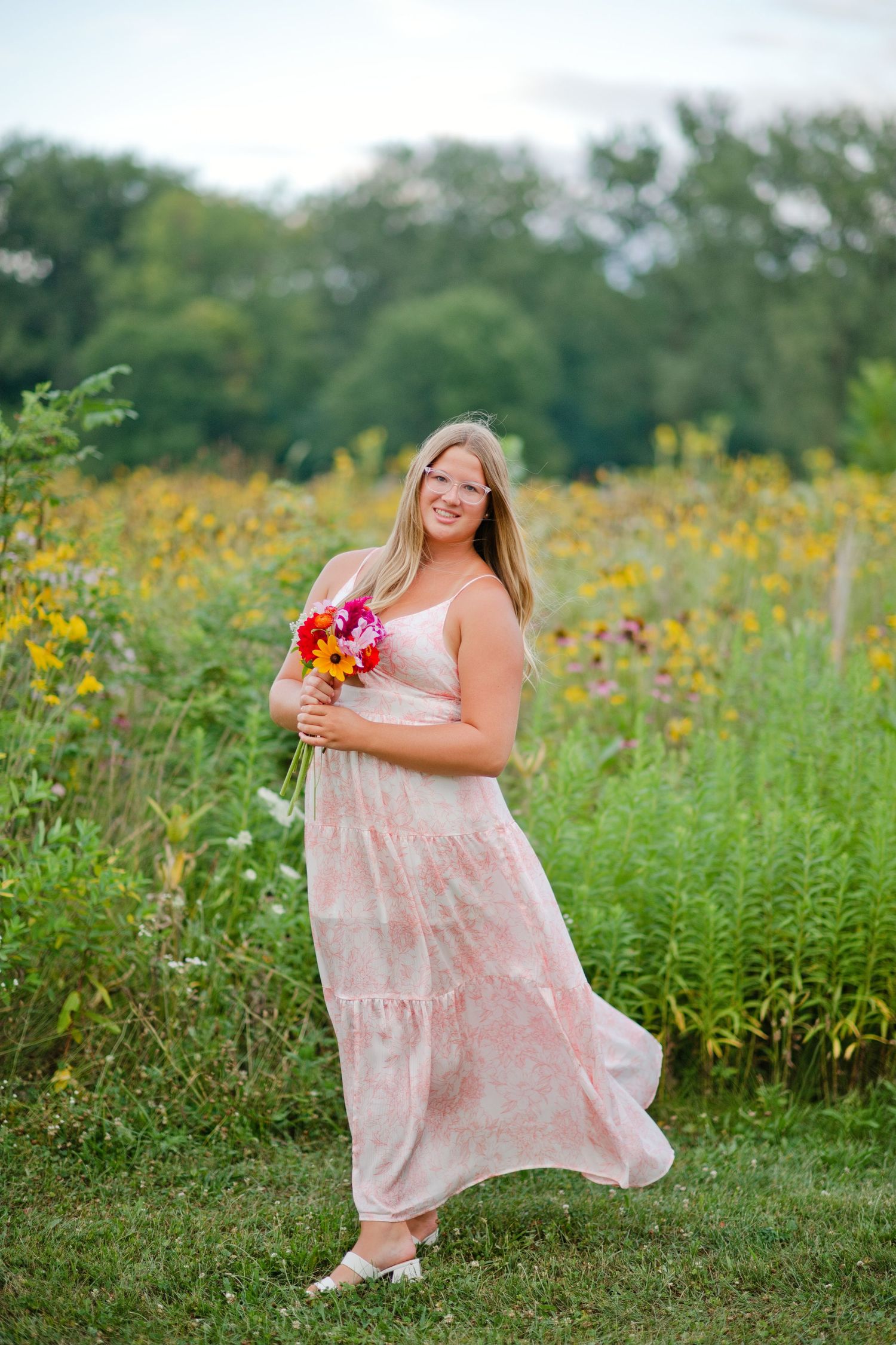 A smiling blonde woman in a flowing pink floral maxi dress holds a bouquet in a wildflower field.
