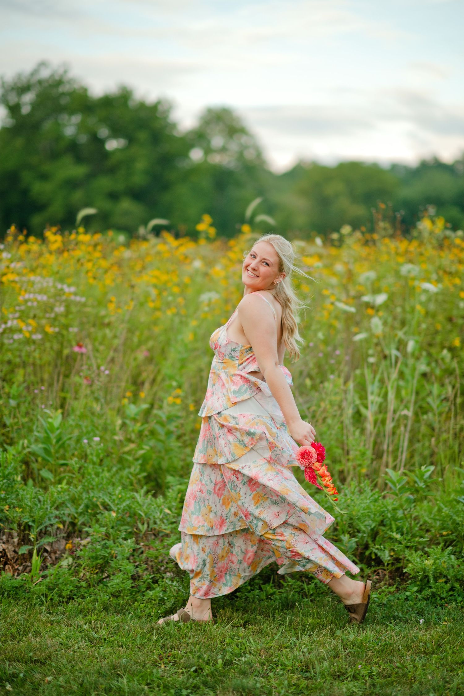 A blonde woman in a flowing floral tiered dress walks through a vibrant wildflower field, smiling over her shoulder.