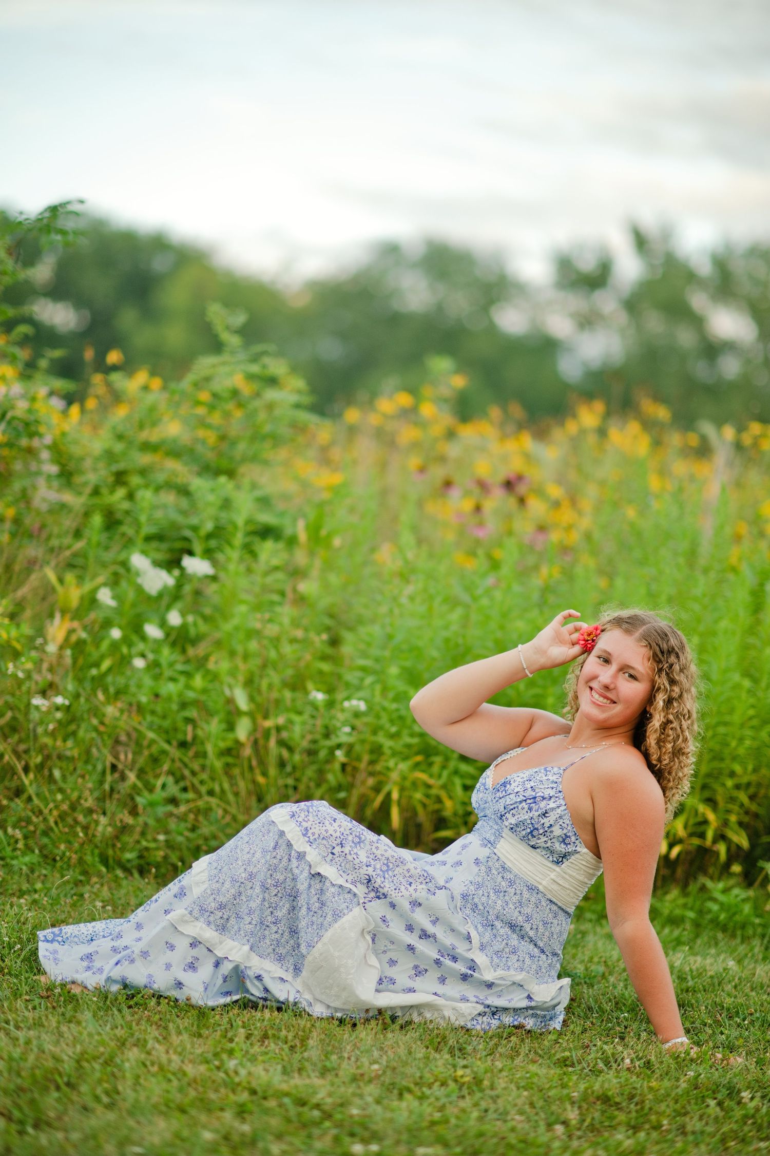 Young woman in blue floral dress sitting on grass in front of wildflower meadow, smiling with flower in hair.