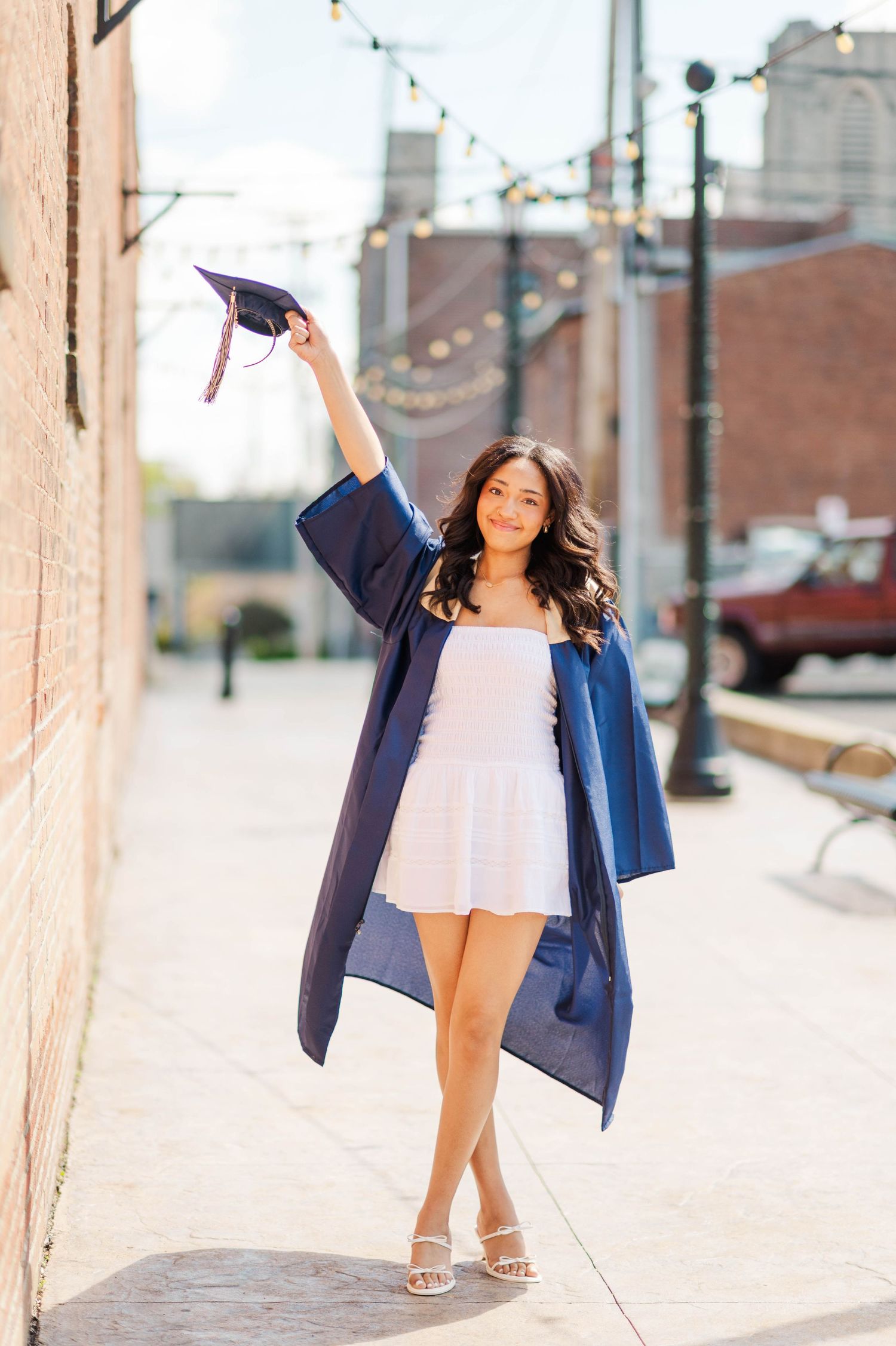 Joyful graduate in navy gown and white dress raises cap triumphantly on sunny urban brick sidewalk.