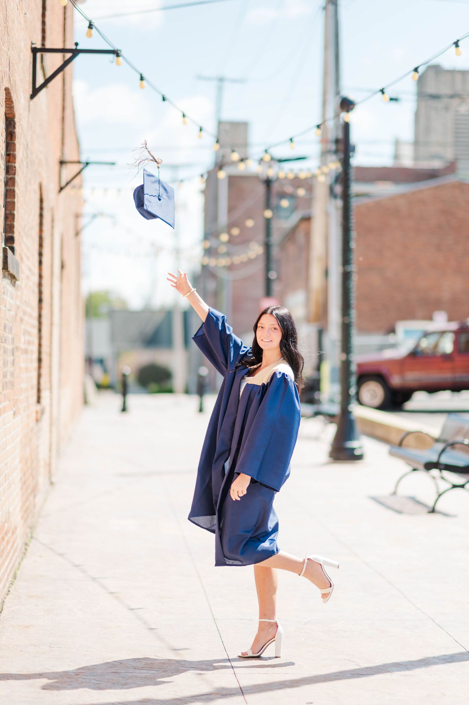 Happy brunette graduate in navy gown tossing mortarboard cap in the air on urban street.