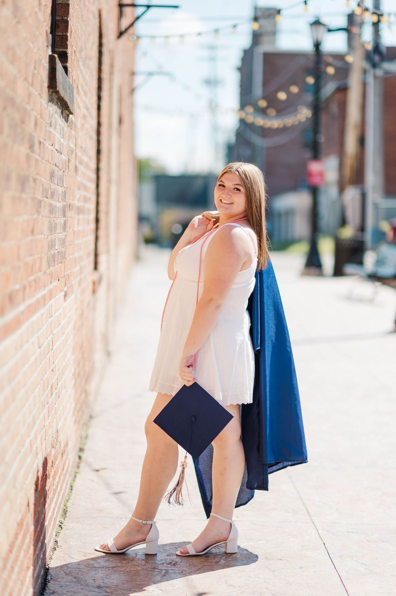 Smiling graduate in white dress holds navy cap while draping graduation gown over shoulder outdoors.