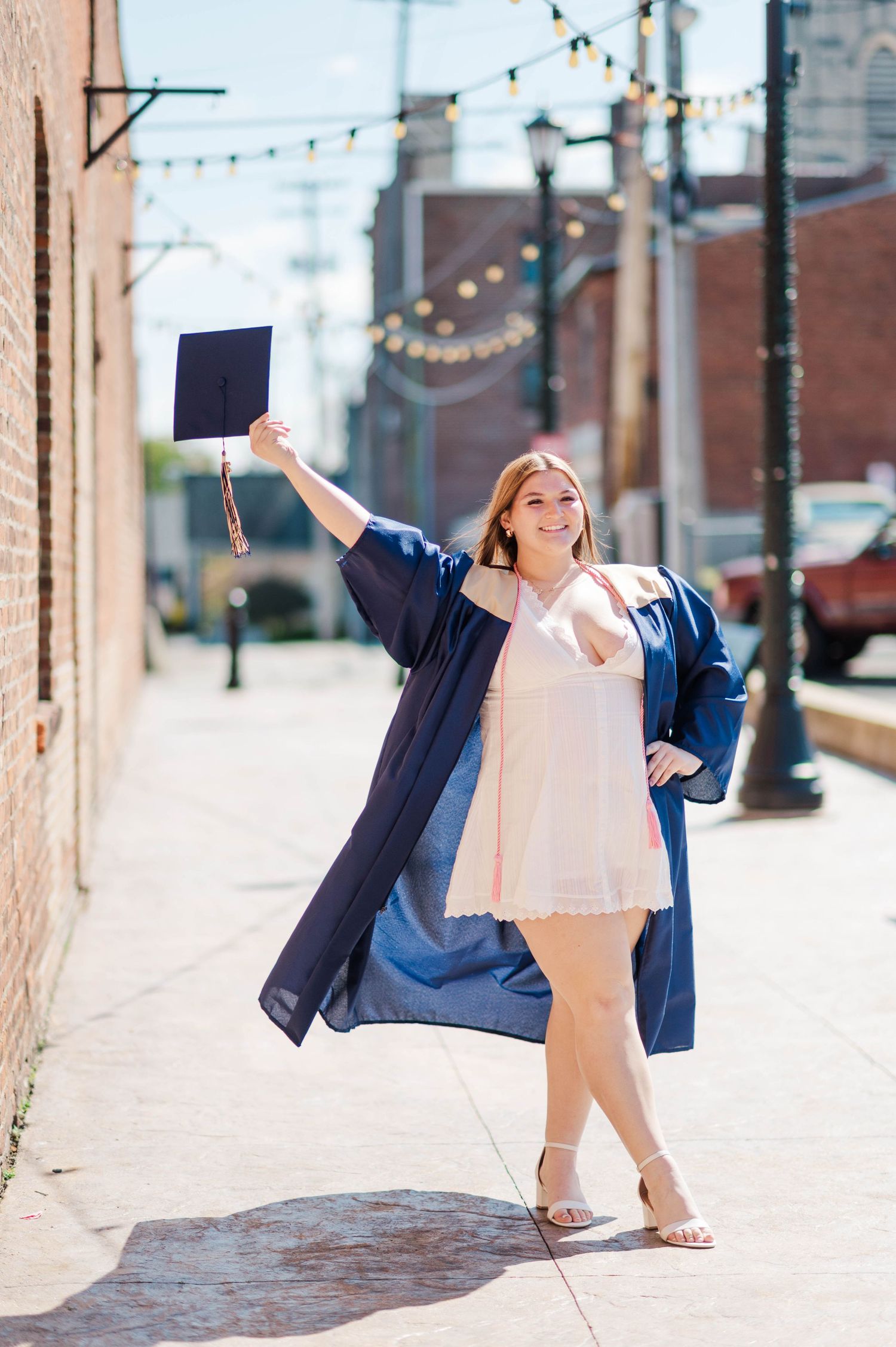 Smiling graduate in navy cap and gown holds mortarboard aloft on sunny urban sidewalk.