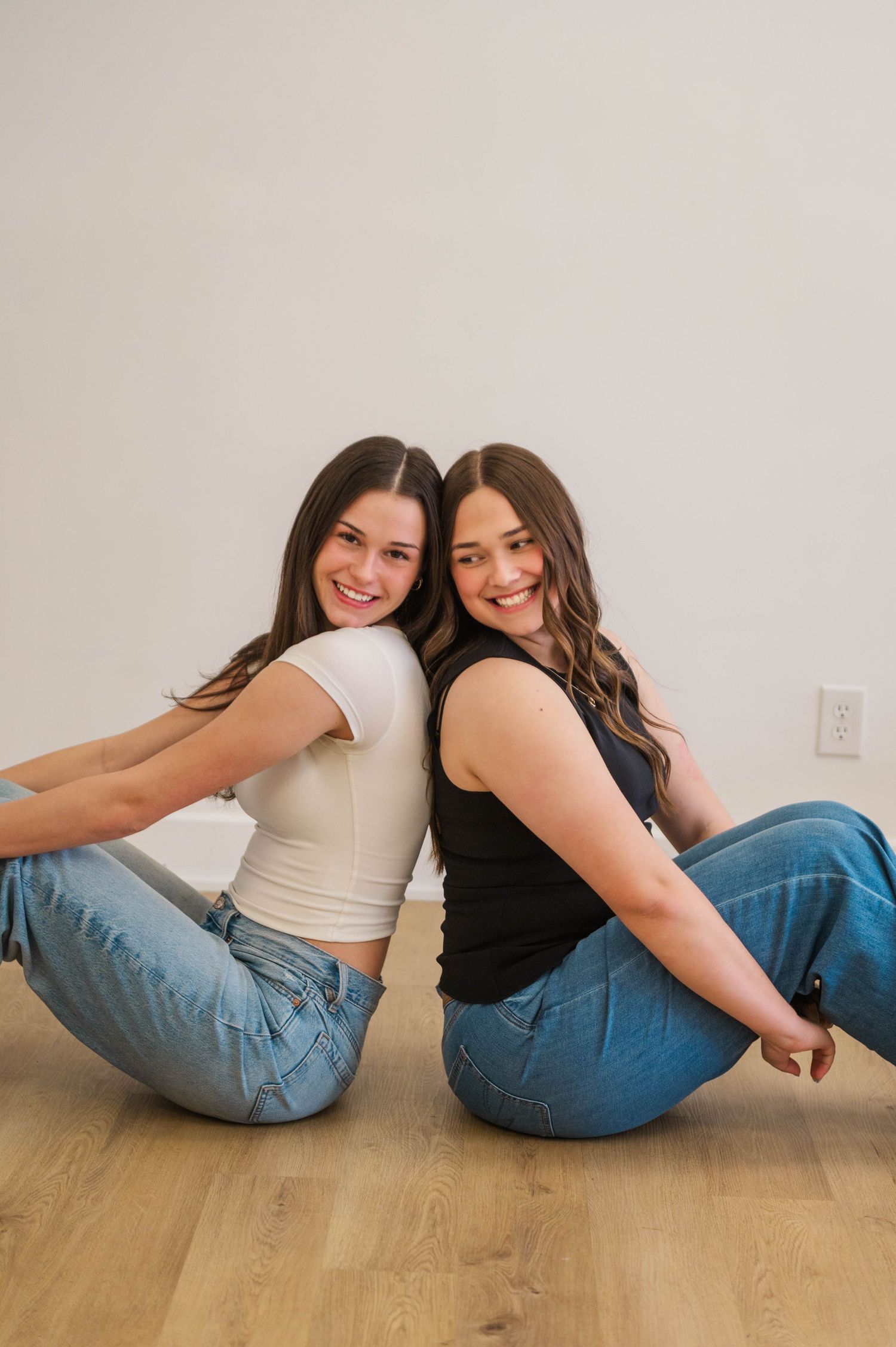 Two brunette women sit back-to-back on hardwood floor smiling, wearing jeans and fitted tops.