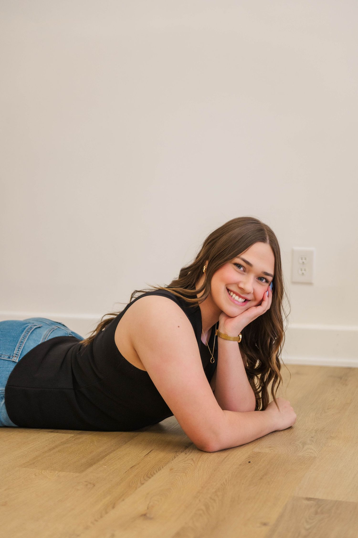 Smiling brunette woman lies on hardwood floor, resting chin on hands, wearing black tank top and blue jeans.