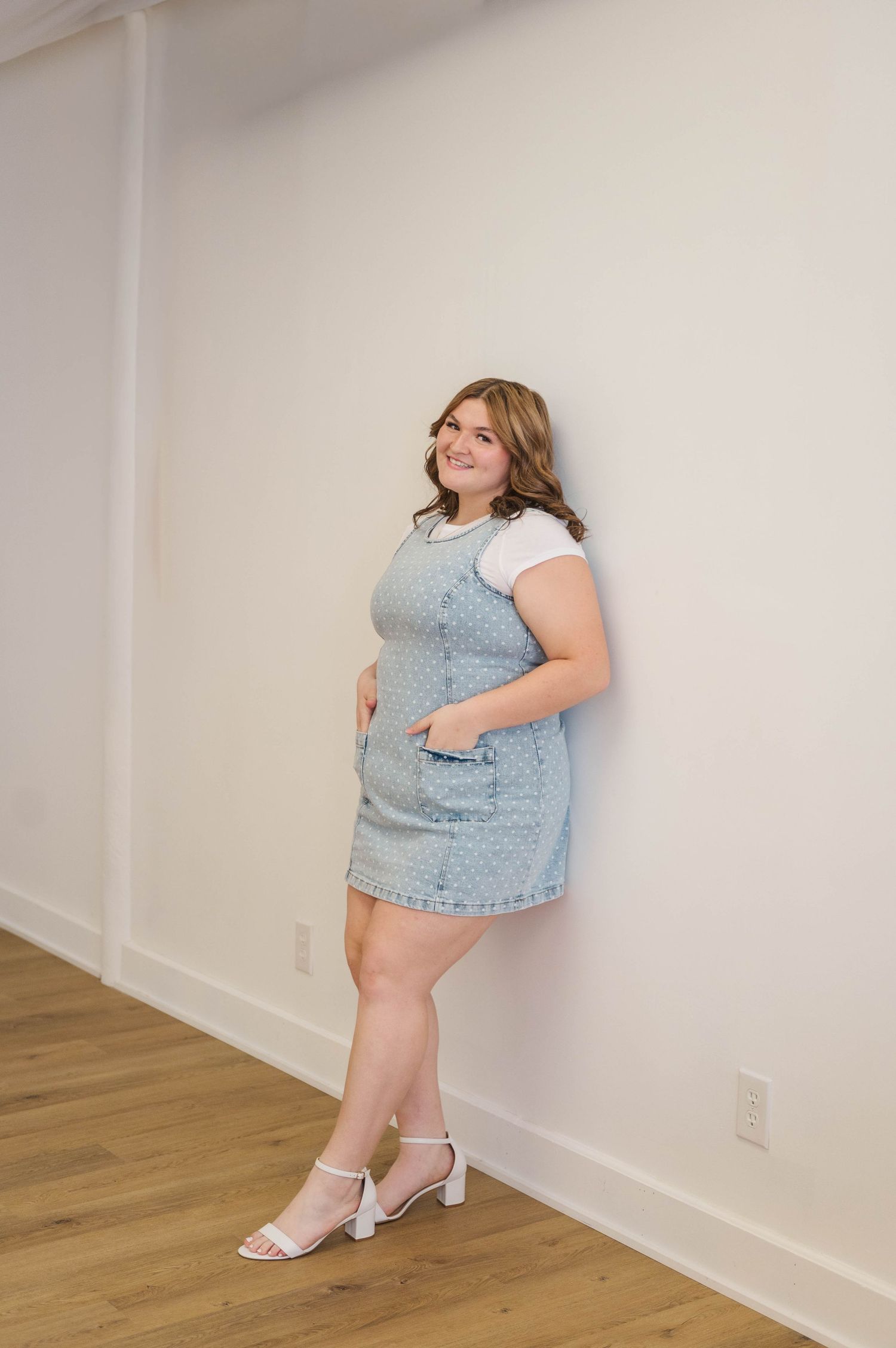 Plus-size woman in denim pinafore dress and white tee leans against white wall in heeled sandals.