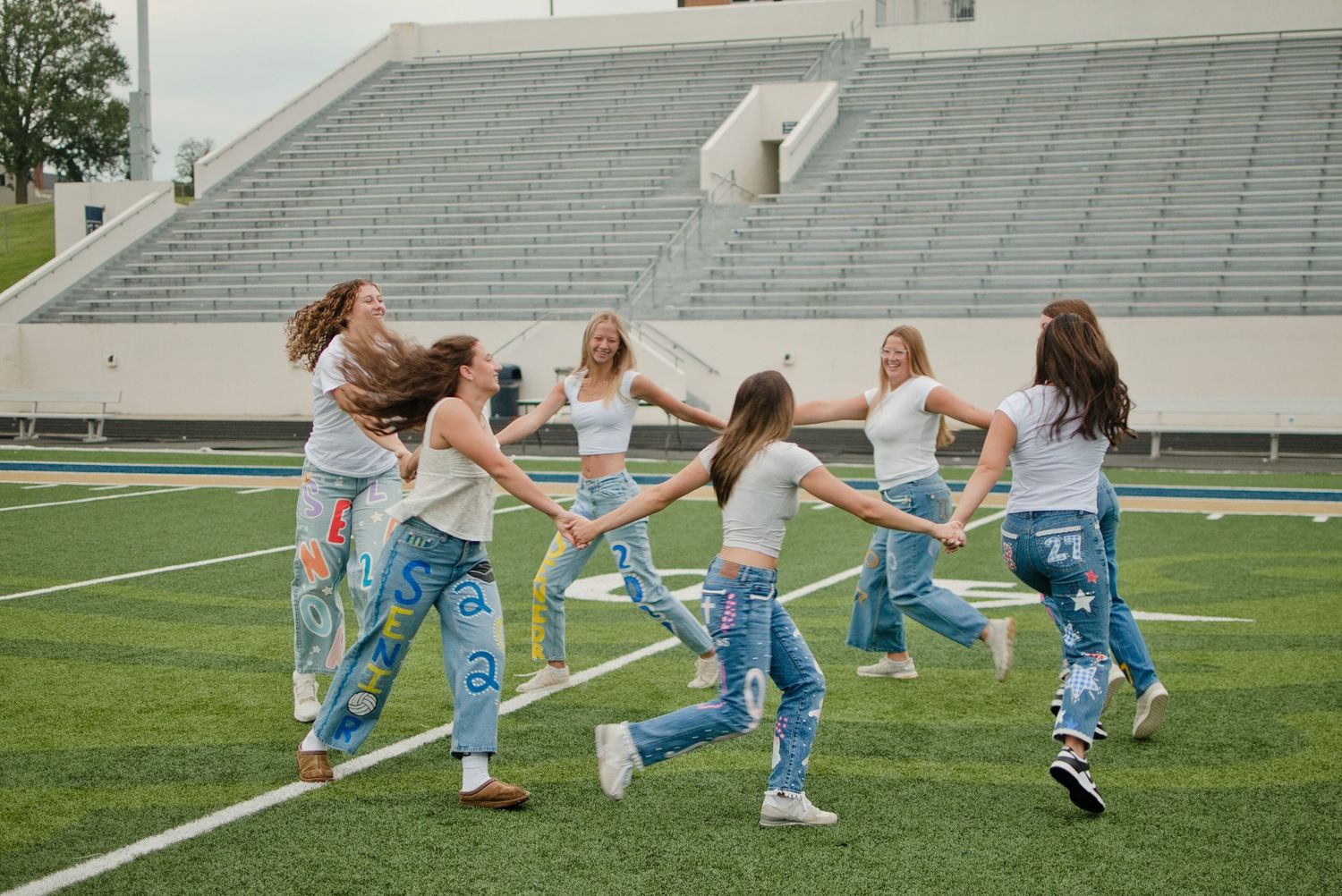 Seven teen girls in white tops and painted senior jeans hold hands in a circle dancing on a football field.