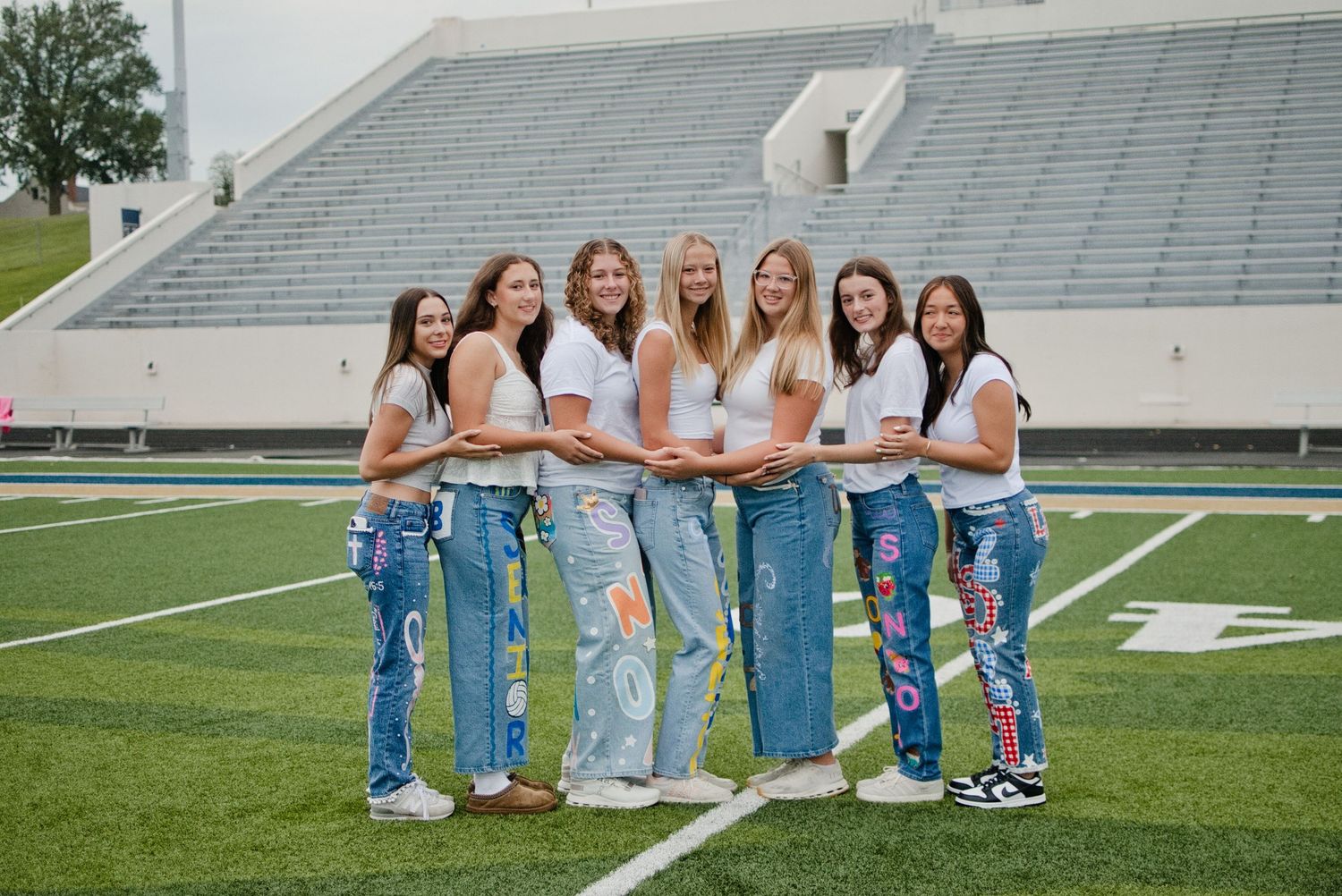 Seven young women in decorated jeans pose together on a football field in front of stadium bleachers.