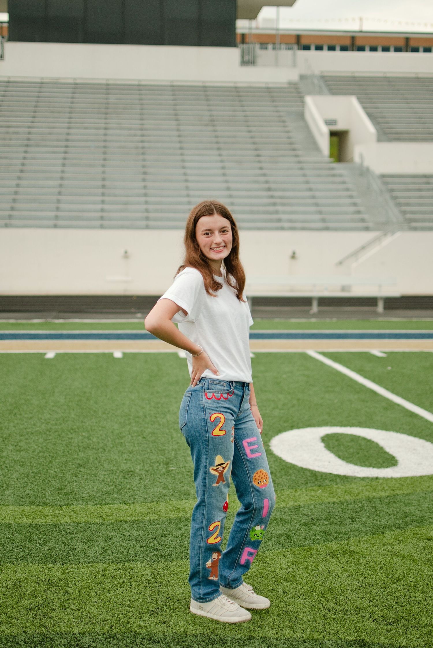 Teen girl posing on football field in white tee and colorful number-decorated jeans with stadium bleachers in background.