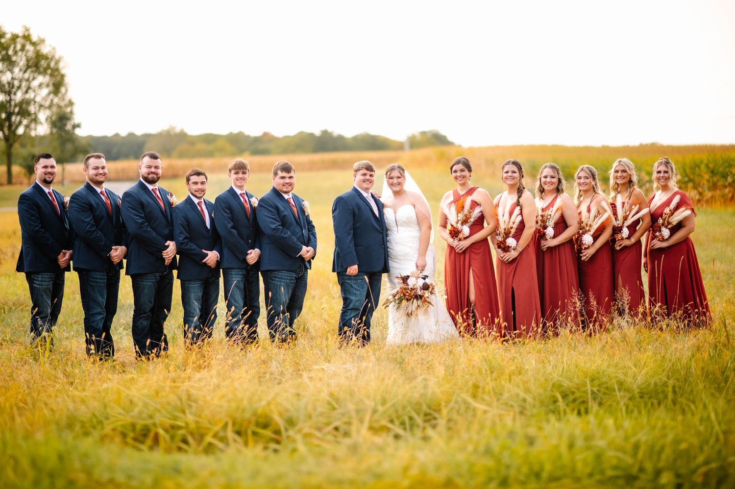 Wedding party lineup with navy suits and red dresses standing in a scenic autumn field.