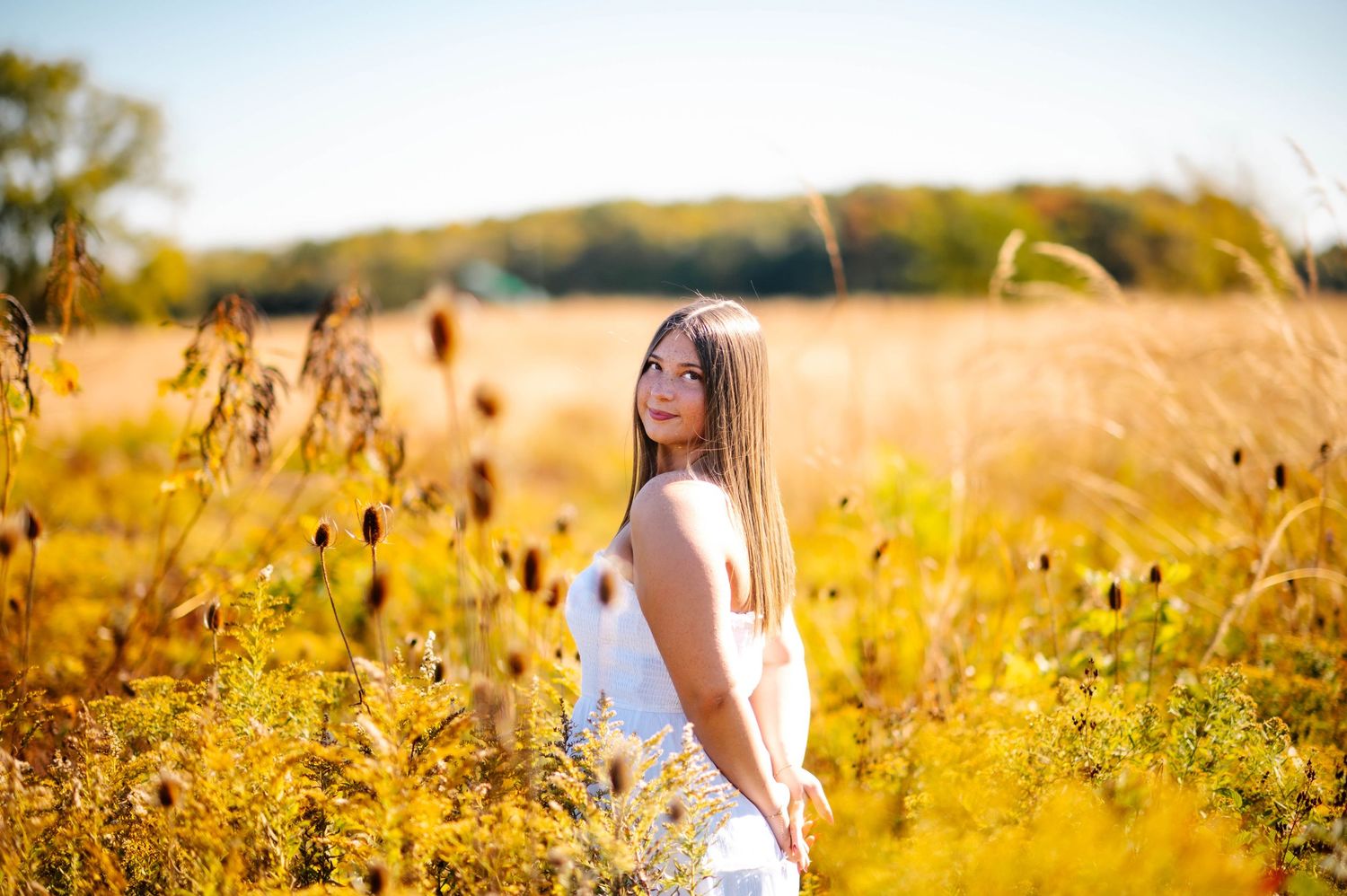 Young woman in white dress standing in golden field with tall wildflowers on a sunny autumn day.