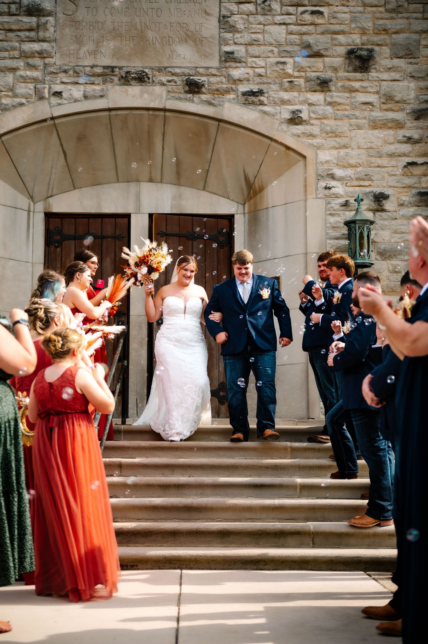 Wedding party in navy suits and coral dresses gather on stone church steps for photos.
