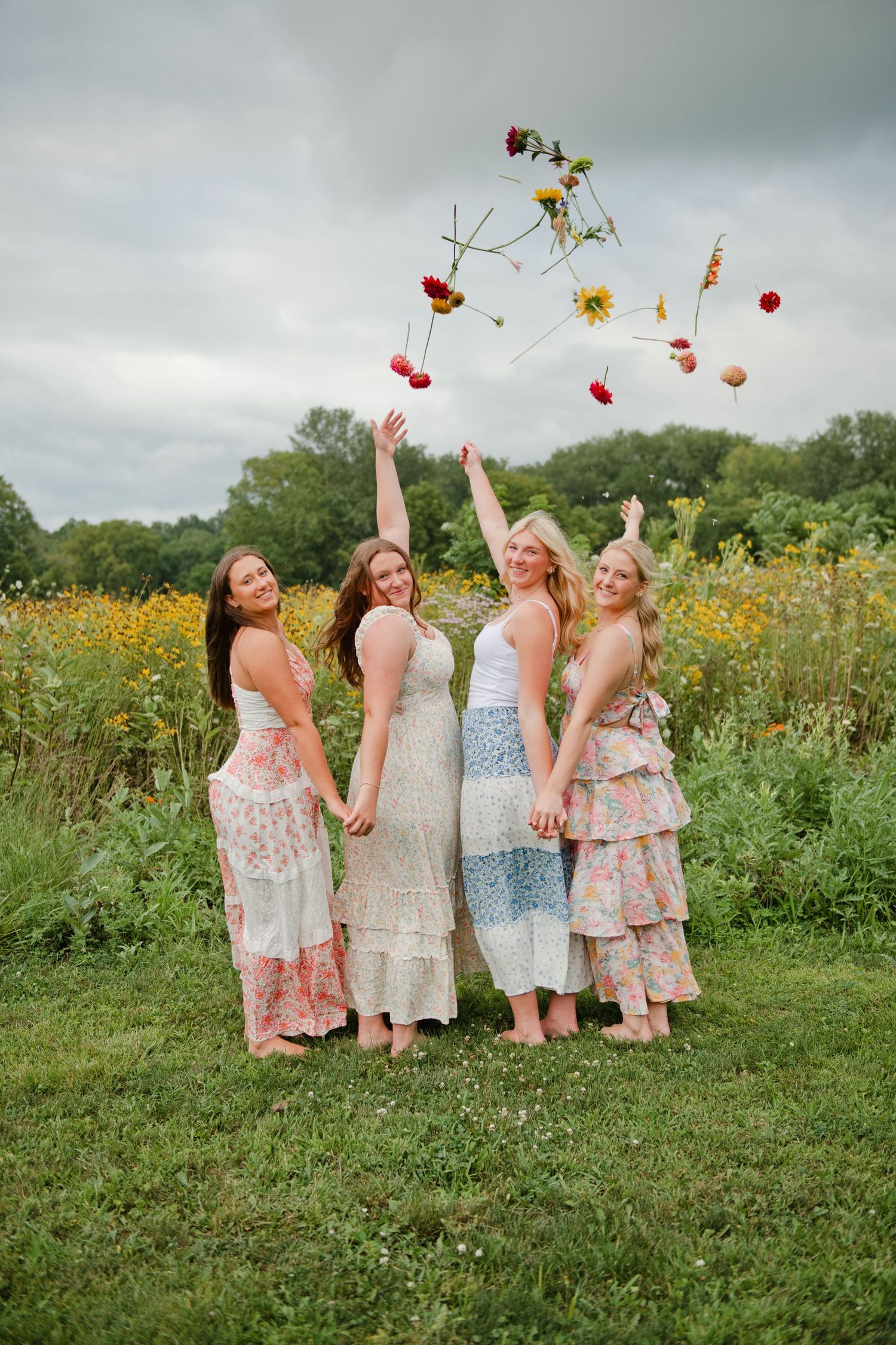 Four women in floral dresses toss wildflowers in the air while holding hands in a lush green meadow.