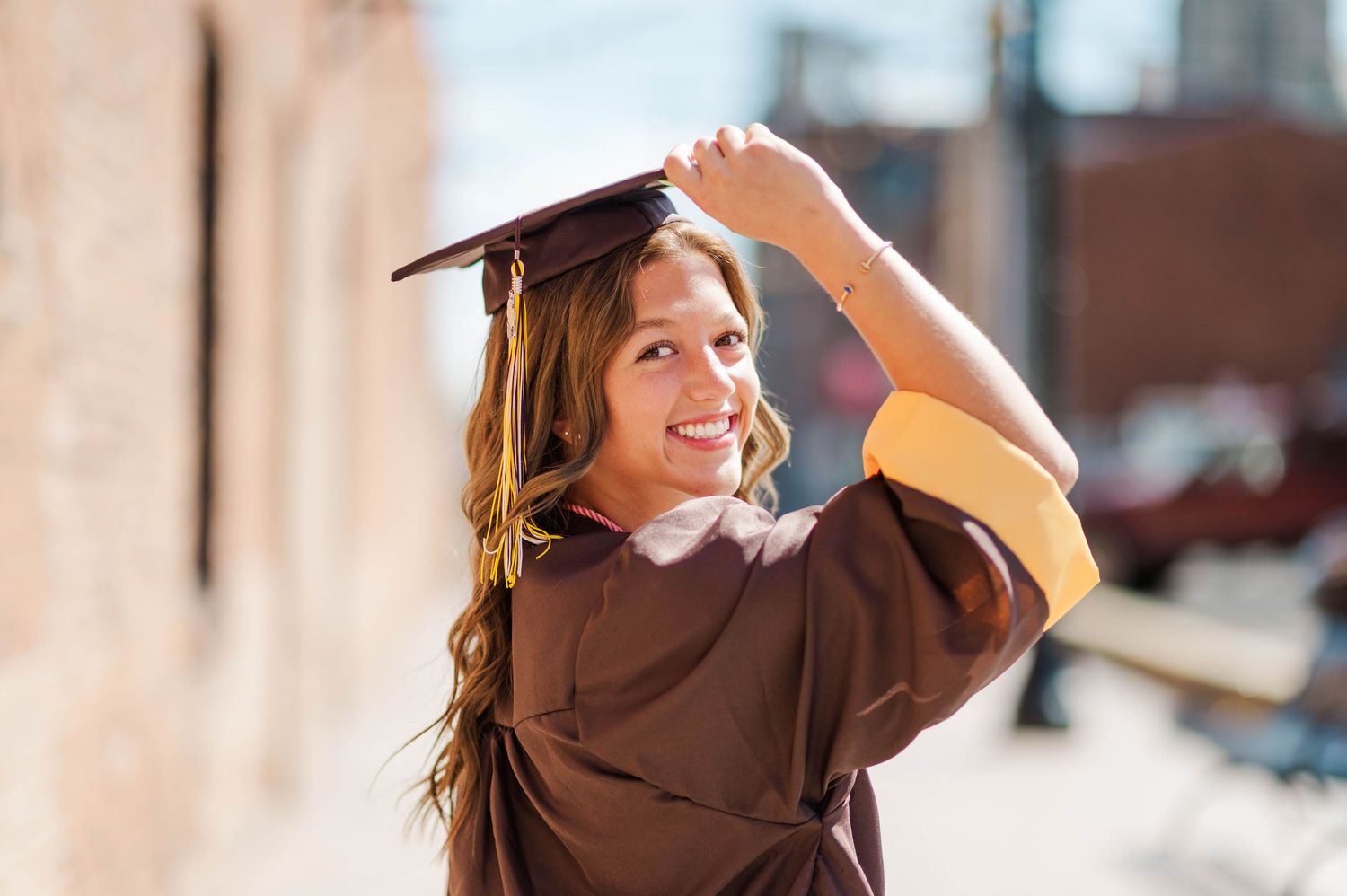 Smiling graduate in brown cap and gown adjusts mortarboard outdoors on sunny day.
