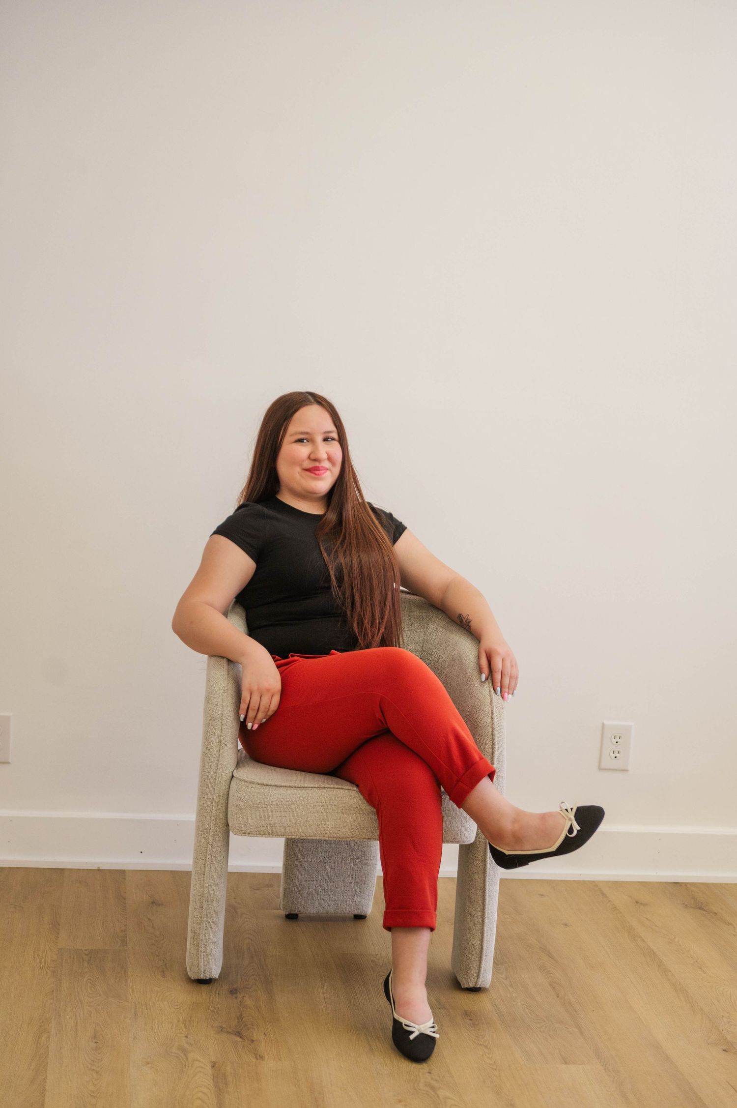 Woman in black top and red pants sitting cross-legged in neutral chair against white wall.