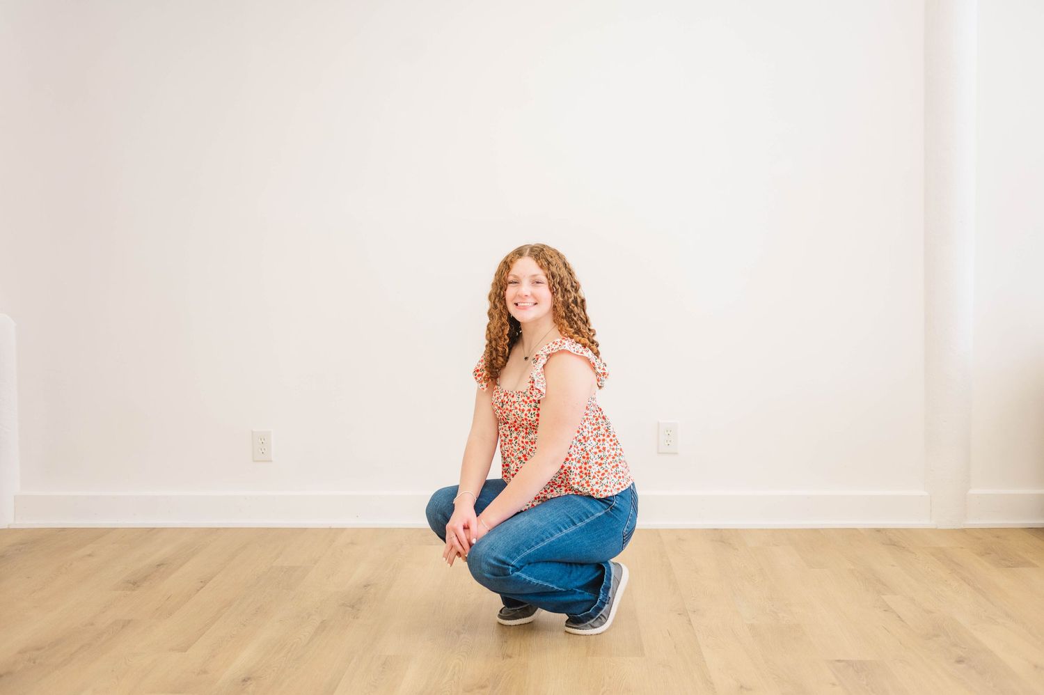Young woman with curly red hair crouching and smiling in a bright white room with light wood floors.
