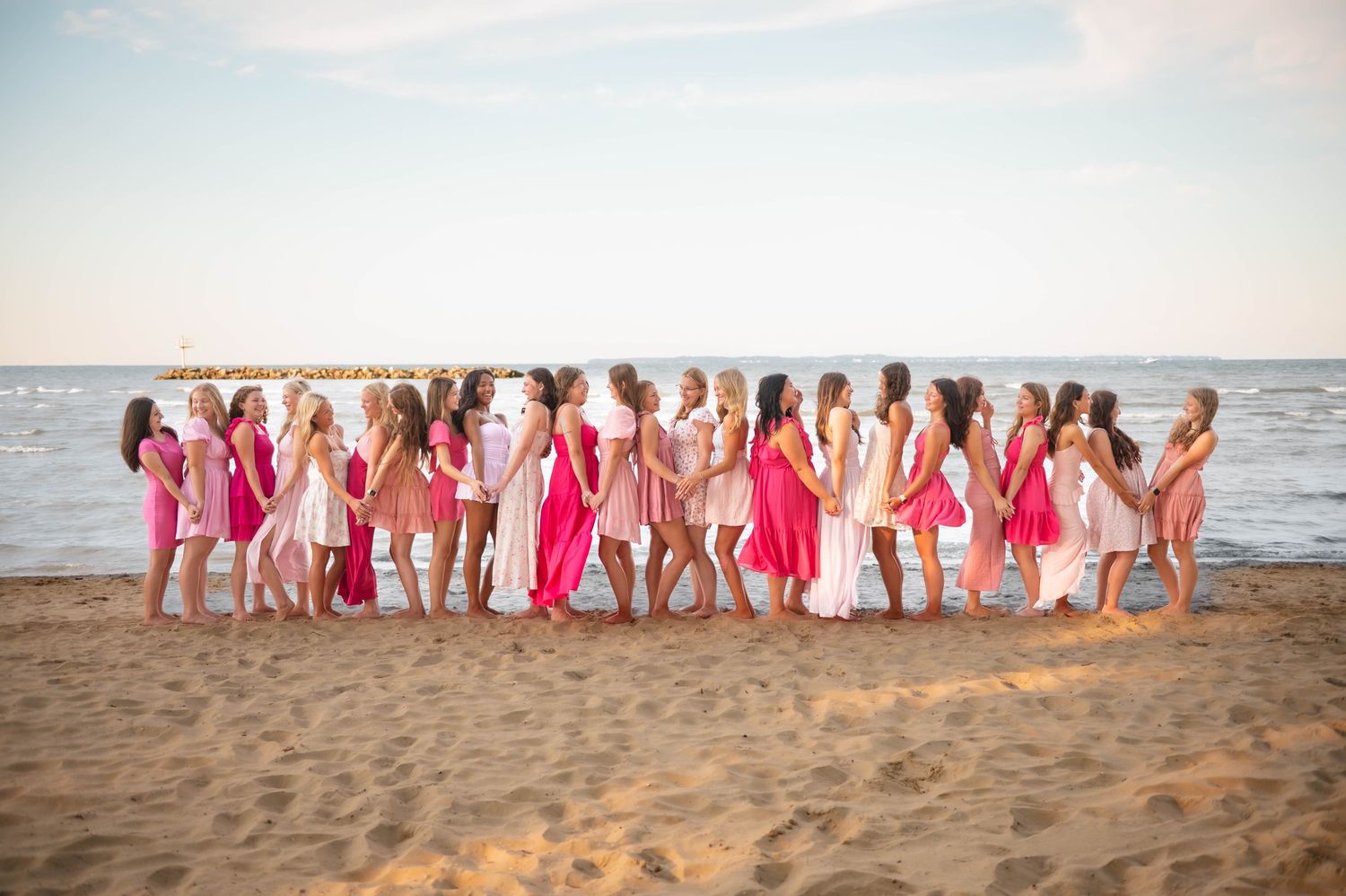 A line of people wearing pink and white dresses stand together on a sandy beach.