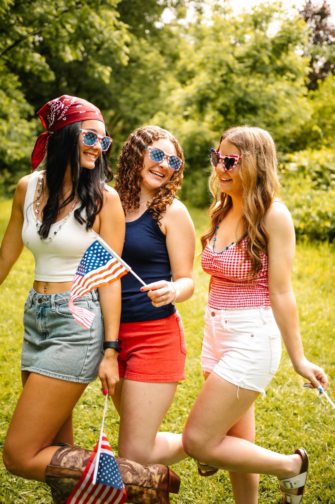 Friends in patriotic summer outfits sitting in grass holding small American flags.