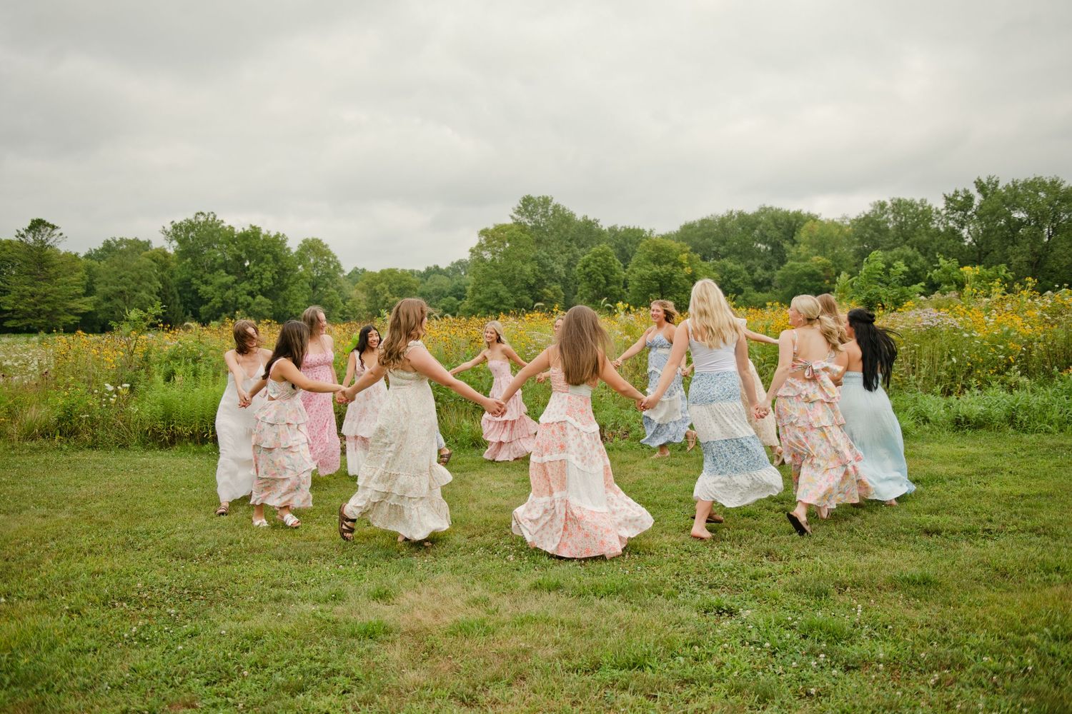 Girls in floral dresses hold hands dancing in a circle on green grass near a wildflower field on a cloudy day.