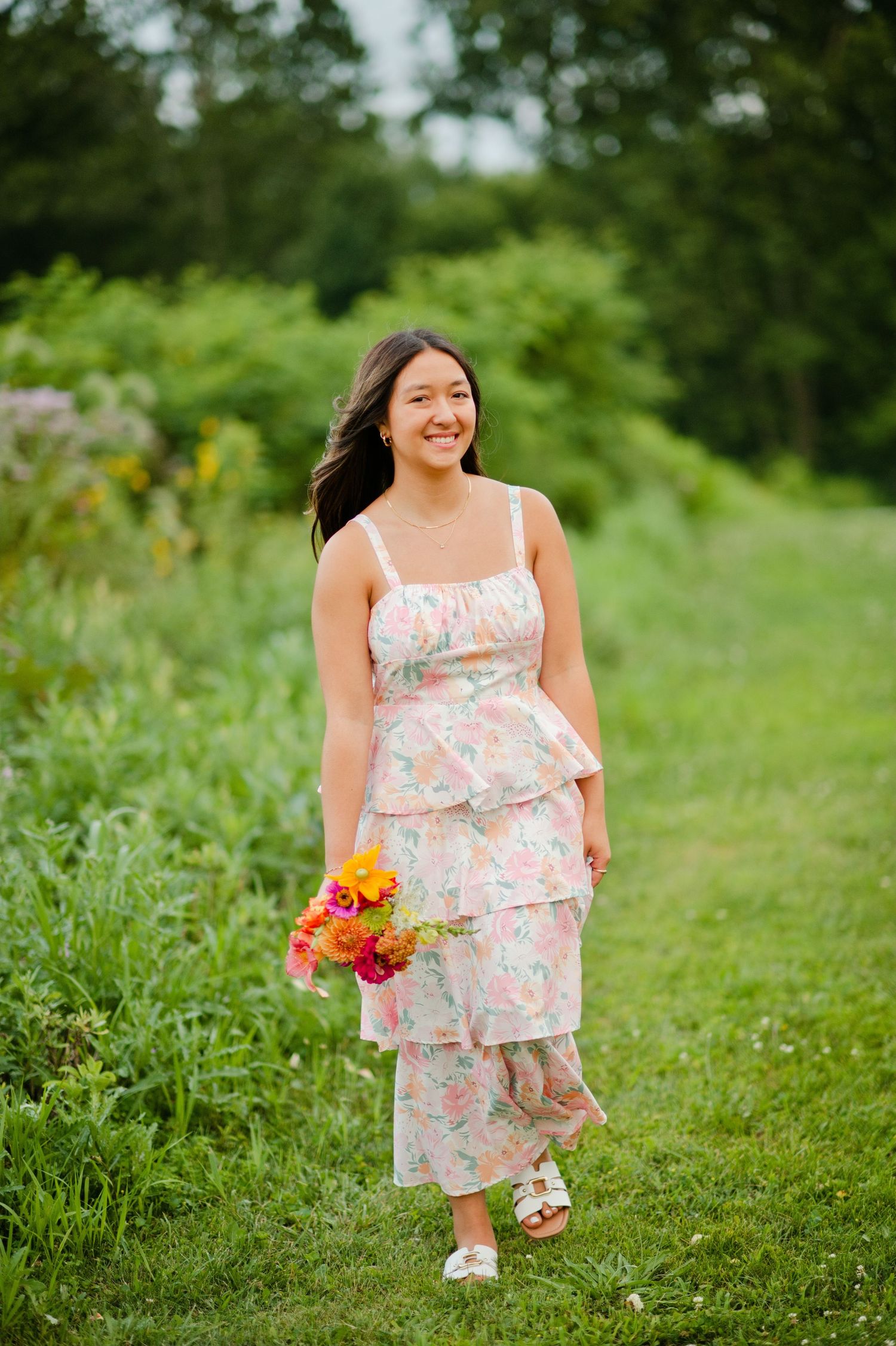 Smiling woman in pastel floral tiered dress walks through green field holding bright bouquet.