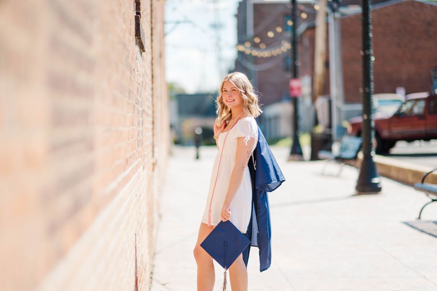 Smiling blonde woman in white dress holding navy blue graduation gown on sunny urban sidewalk.