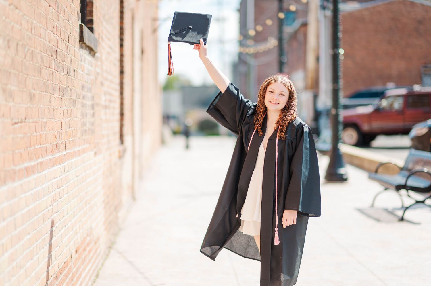 Graduate in black cap and gown raising cap triumphantly beside brick wall outdoors.