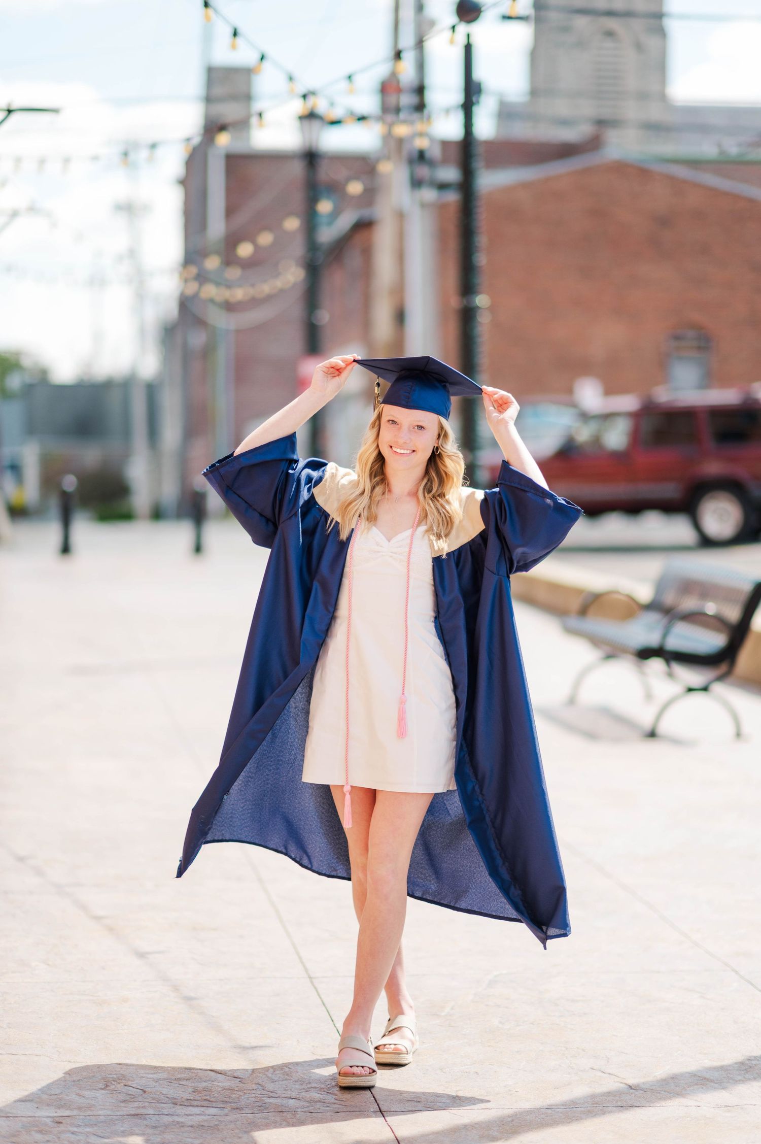 Smiling blonde graduate in navy cap and gown adjusting mortarboard on sunny urban sidewalk.