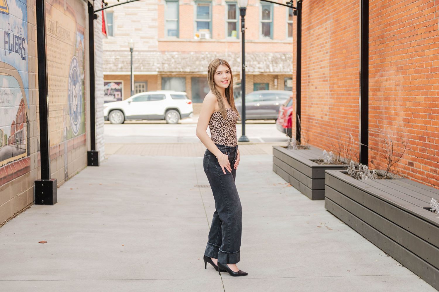 Young woman in leopard top and gray pants poses on urban brick alley sidewalk.