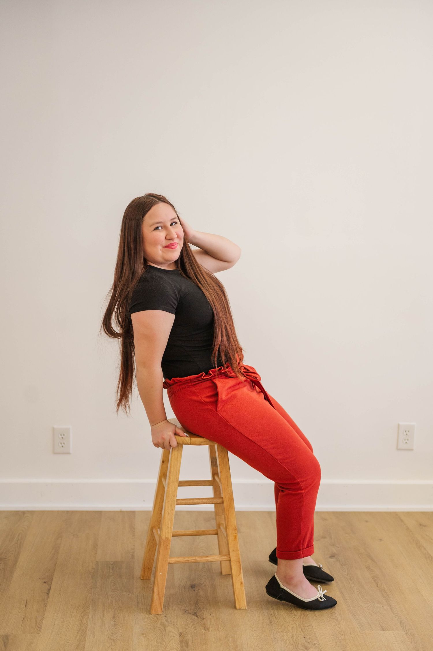Woman with long brown hair sits on wooden stool in black top and red pants, smiling over shoulder.