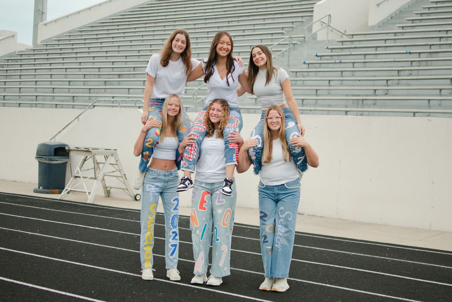 Six teenage girls pose in jeans and white tees on a track field with stadium bleachers in the background.
