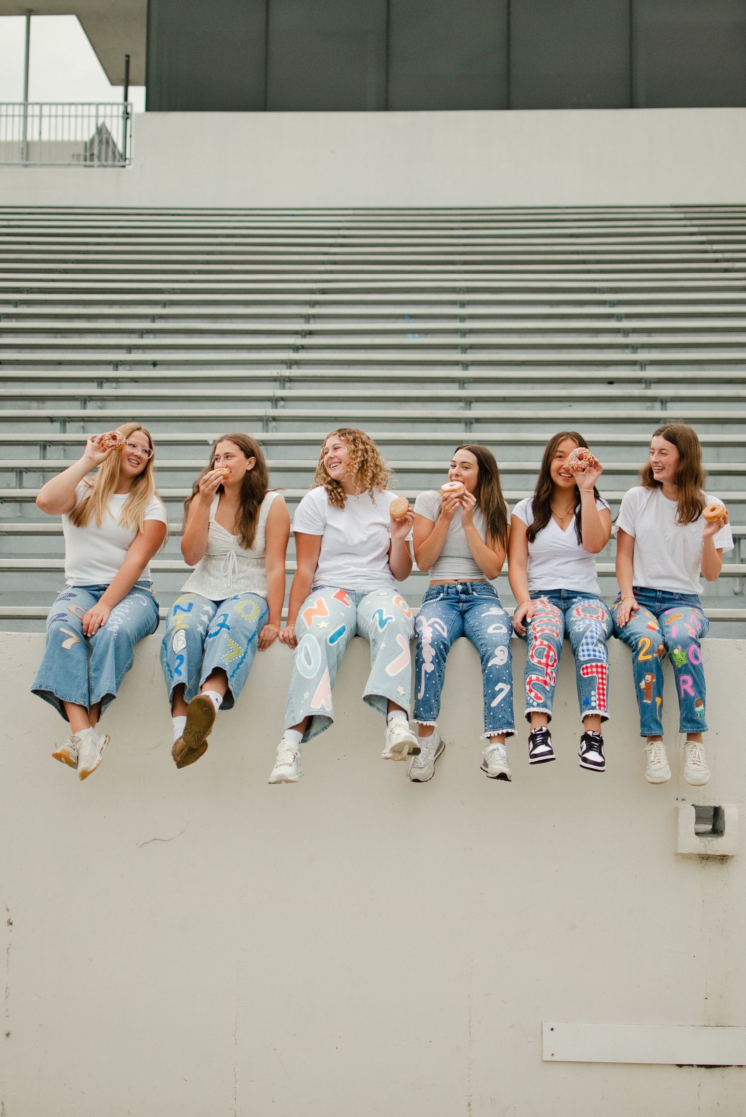 Six girls in white tees and painted senior jeans sit on stadium bleachers eating donuts together.