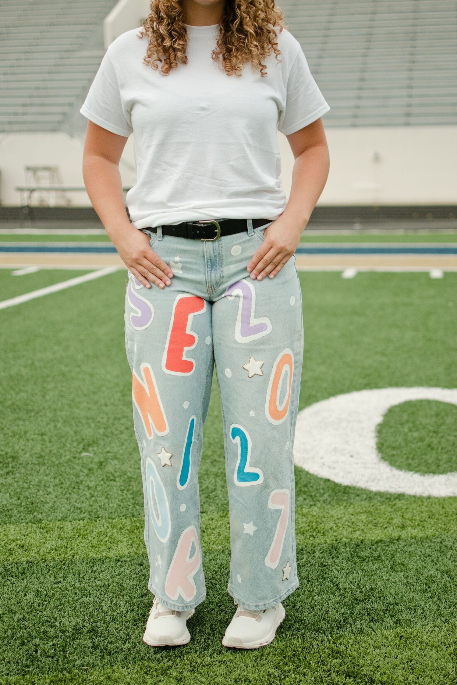 Teen girl wearing colorful letter-painted light wash jeans with stars on a football field.