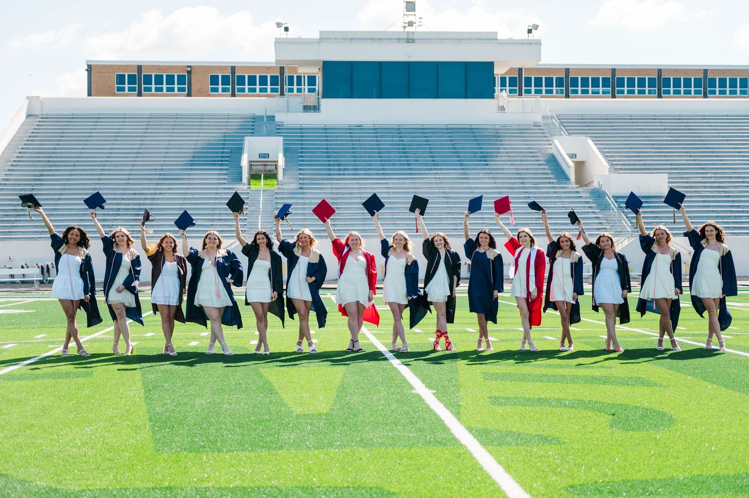 Graduates in caps and gowns toss mortarboards in the air on a football field stadium.