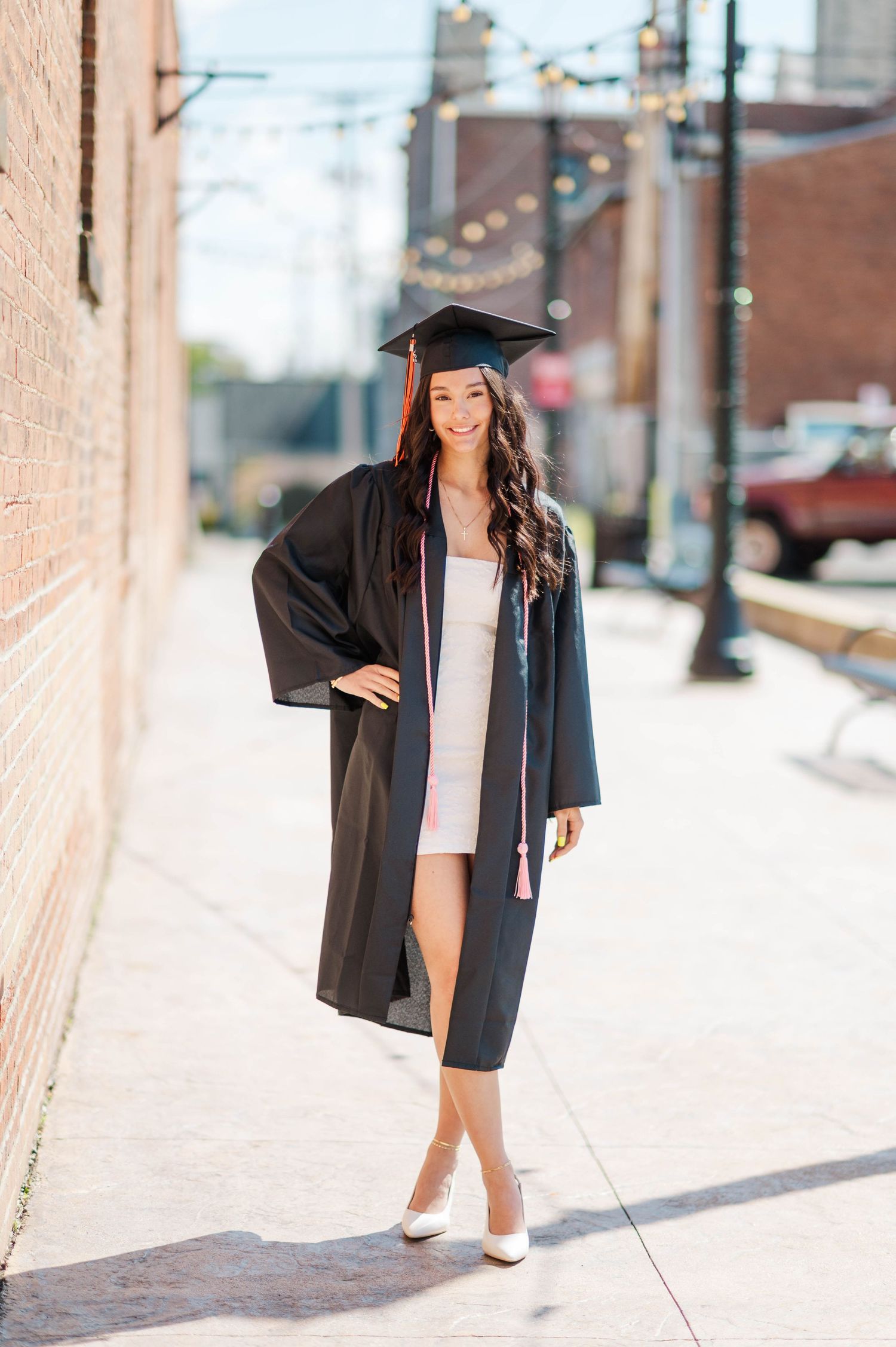 Brunette graduate in black cap and gown with pink tassel posing on sunny urban sidewalk.