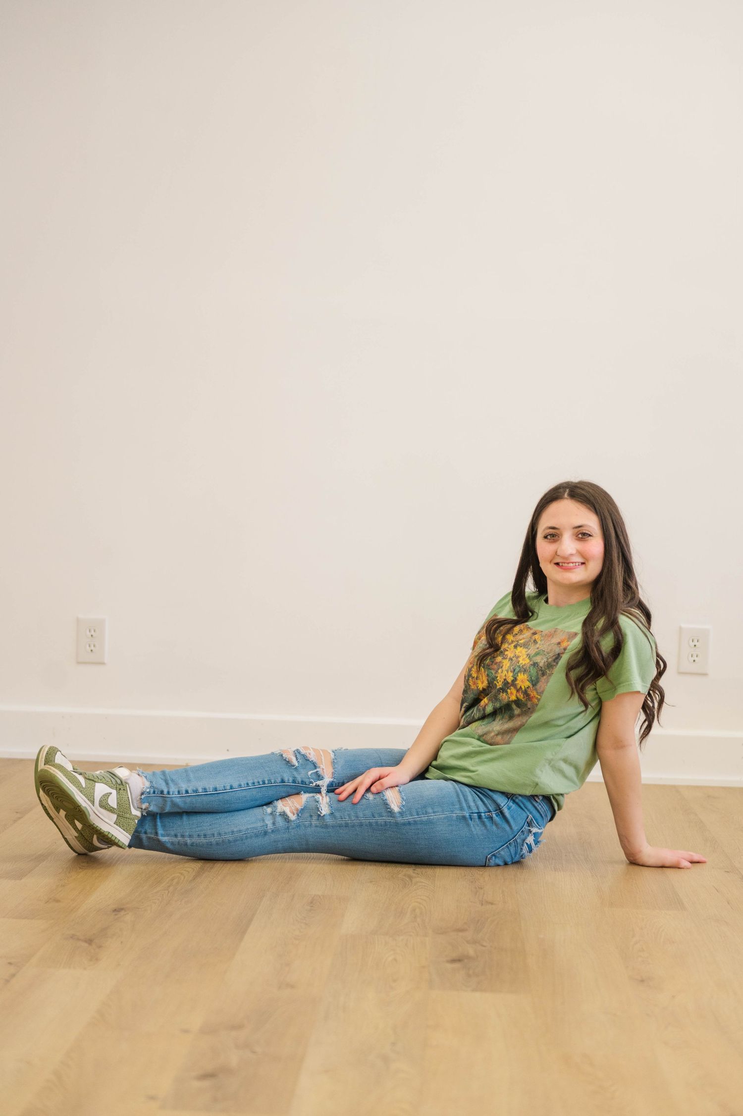Smiling young woman in green graphic tee and ripped jeans sitting casually on hardwood floor.