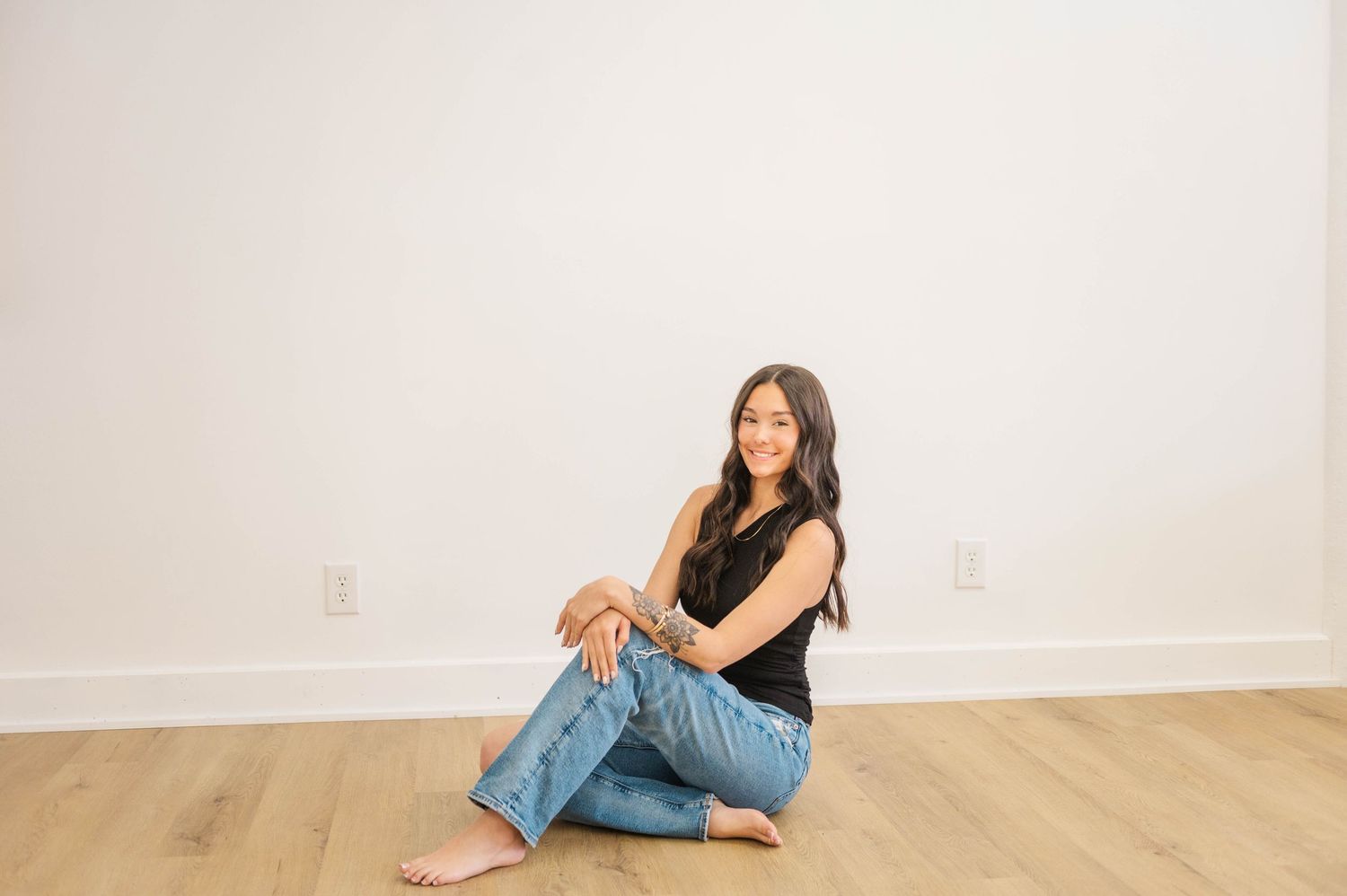 Young woman in black top and jeans sitting cross-legged on hardwood floor, smiling against white wall.
