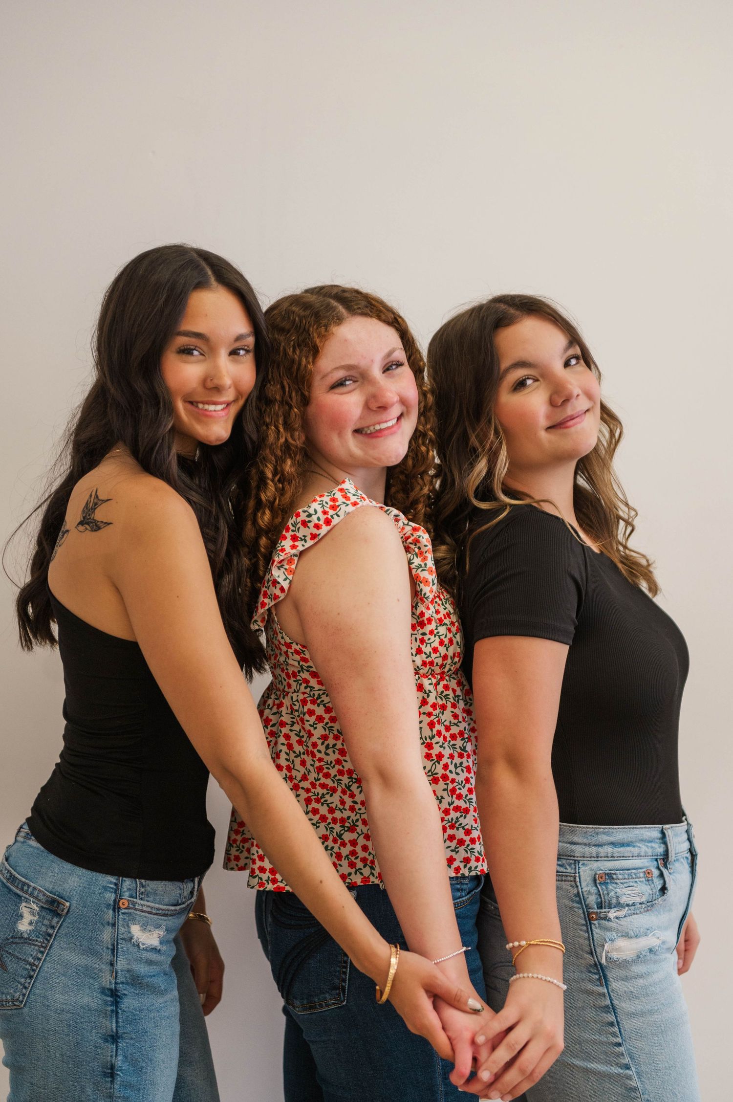 Three young women pose together smiling, wearing casual tops and jeans against a white background.