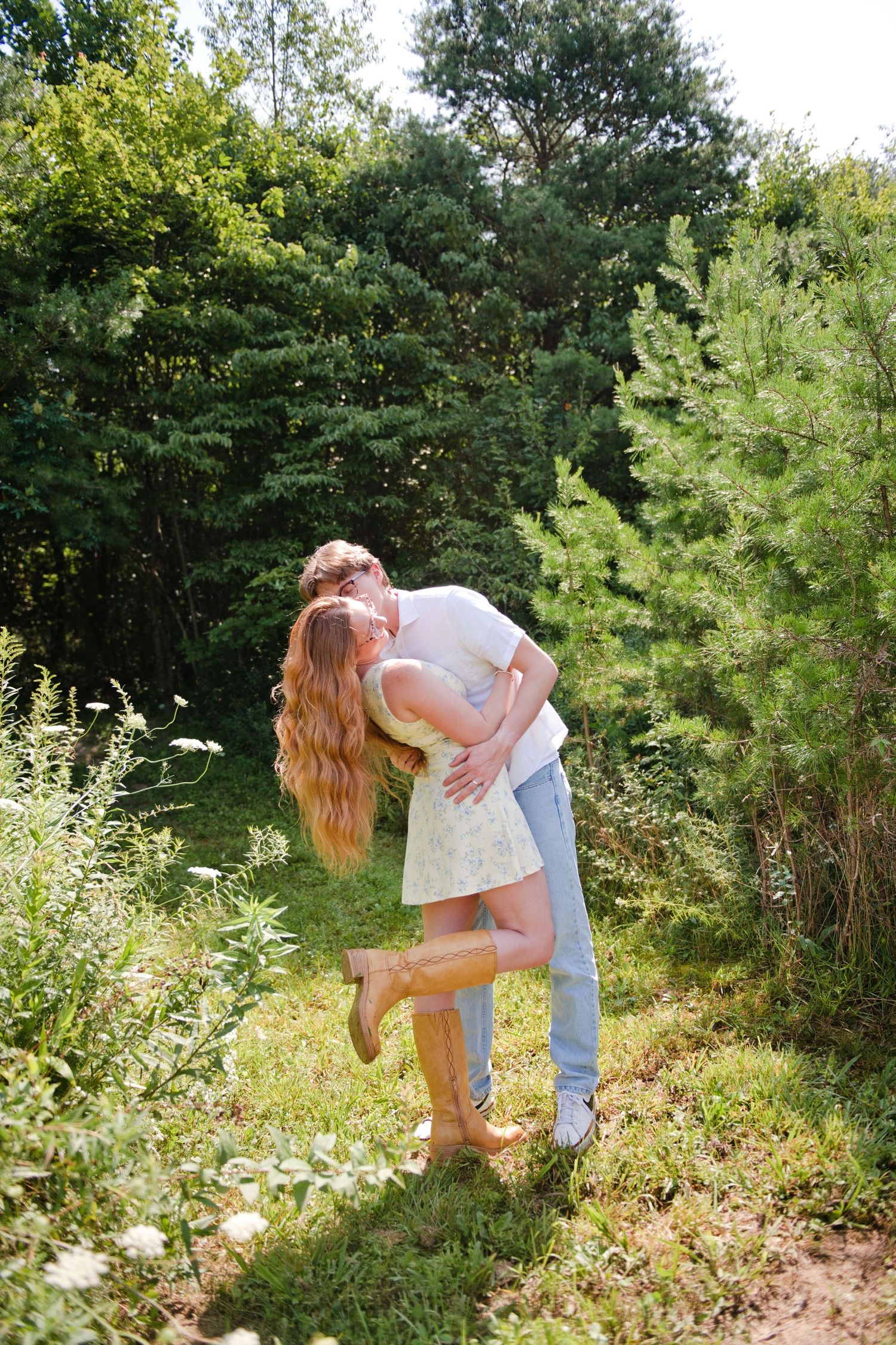 Romantic couple sharing a kiss in a lush green garden, woman in yellow dress lifting her leg joyfully.