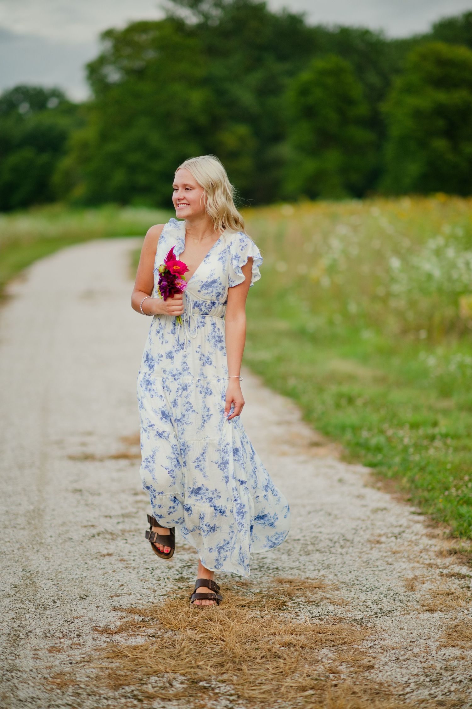 A smiling blonde woman in a blue floral maxi dress walks down a gravel path holding a pink bouquet.